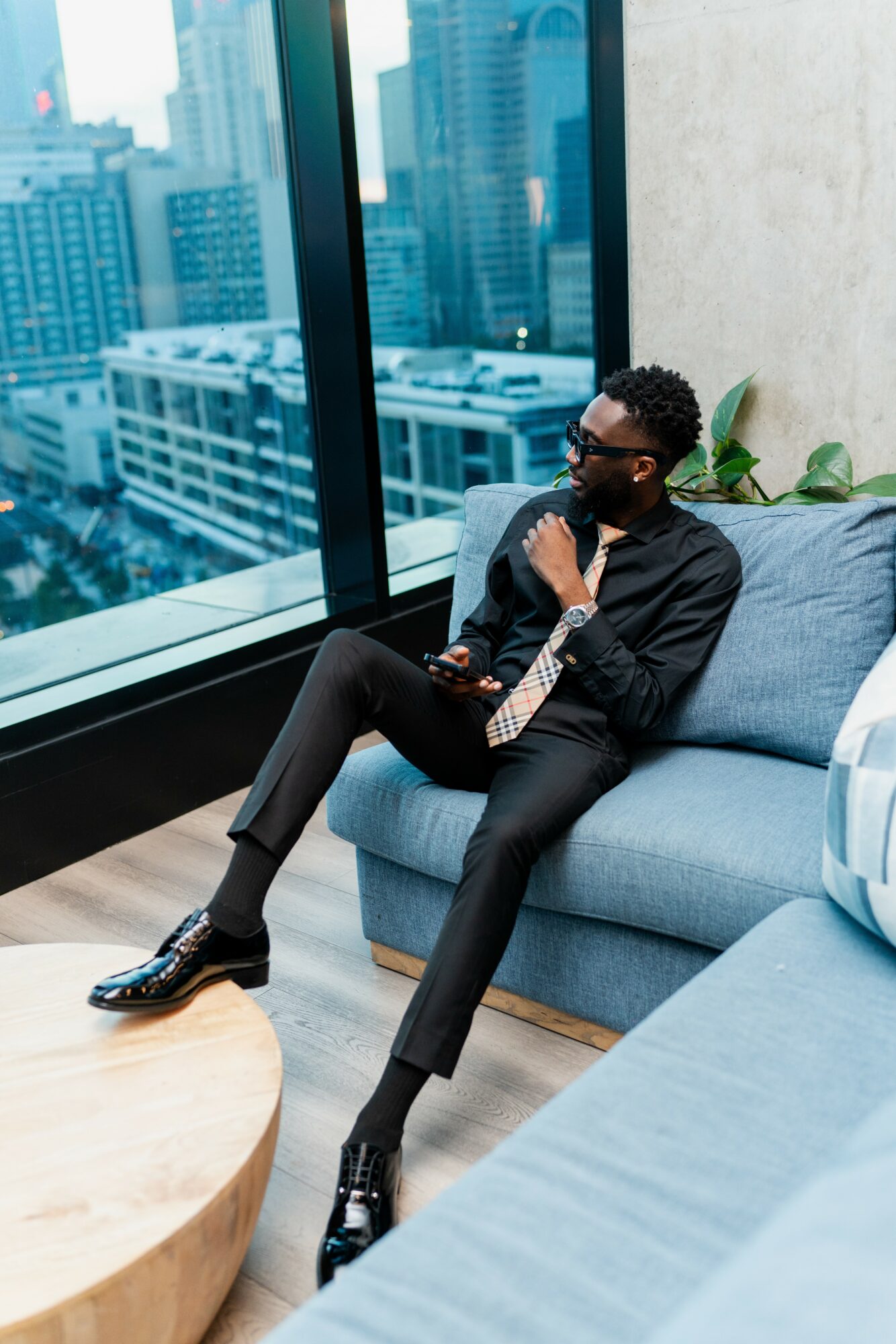 Man with sunglasses in a suit sitting on a blue sofa near large windows overlooking city buildings.