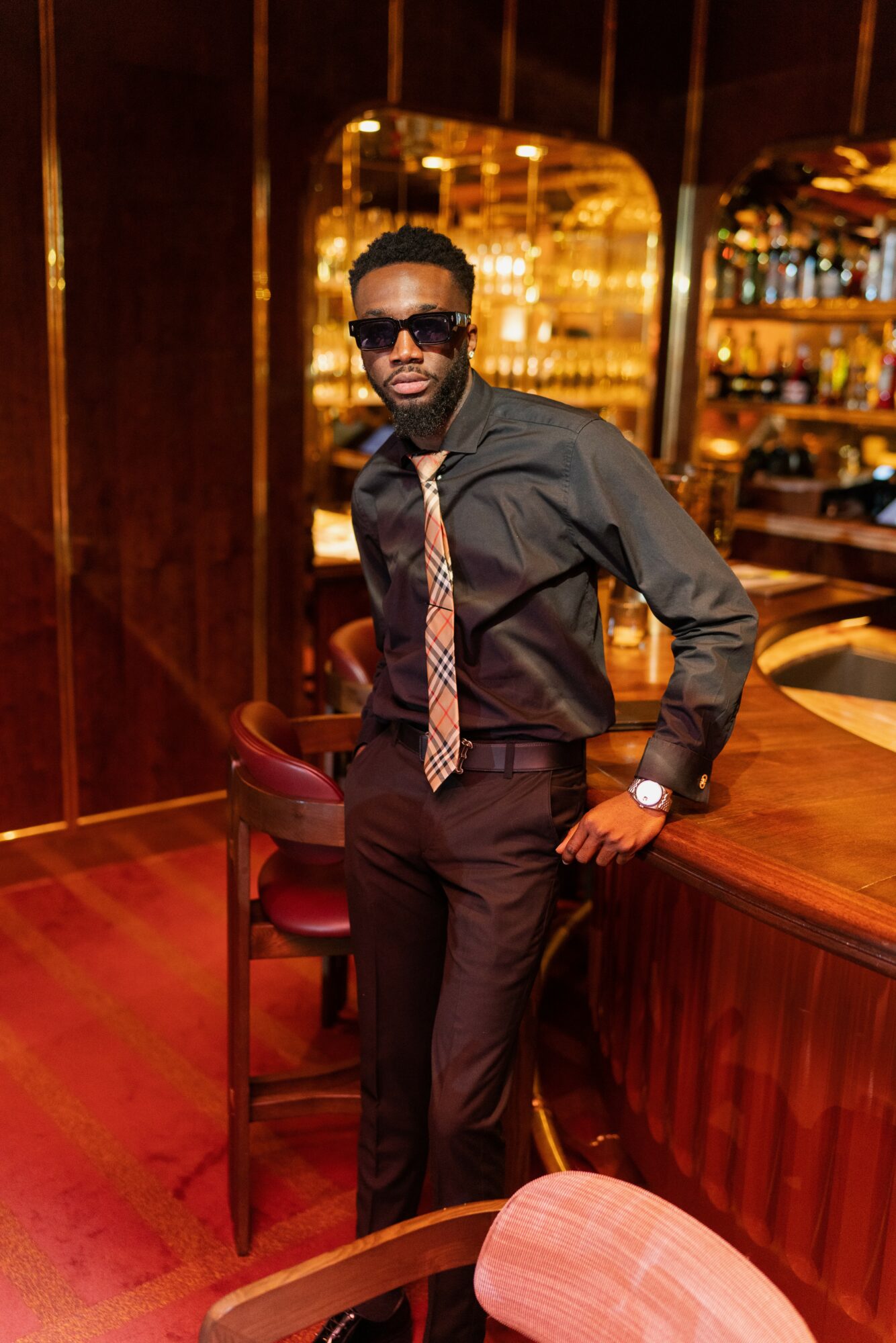 Man in black shirt and tie with sunglasses standing at bar in dimly lit lounge