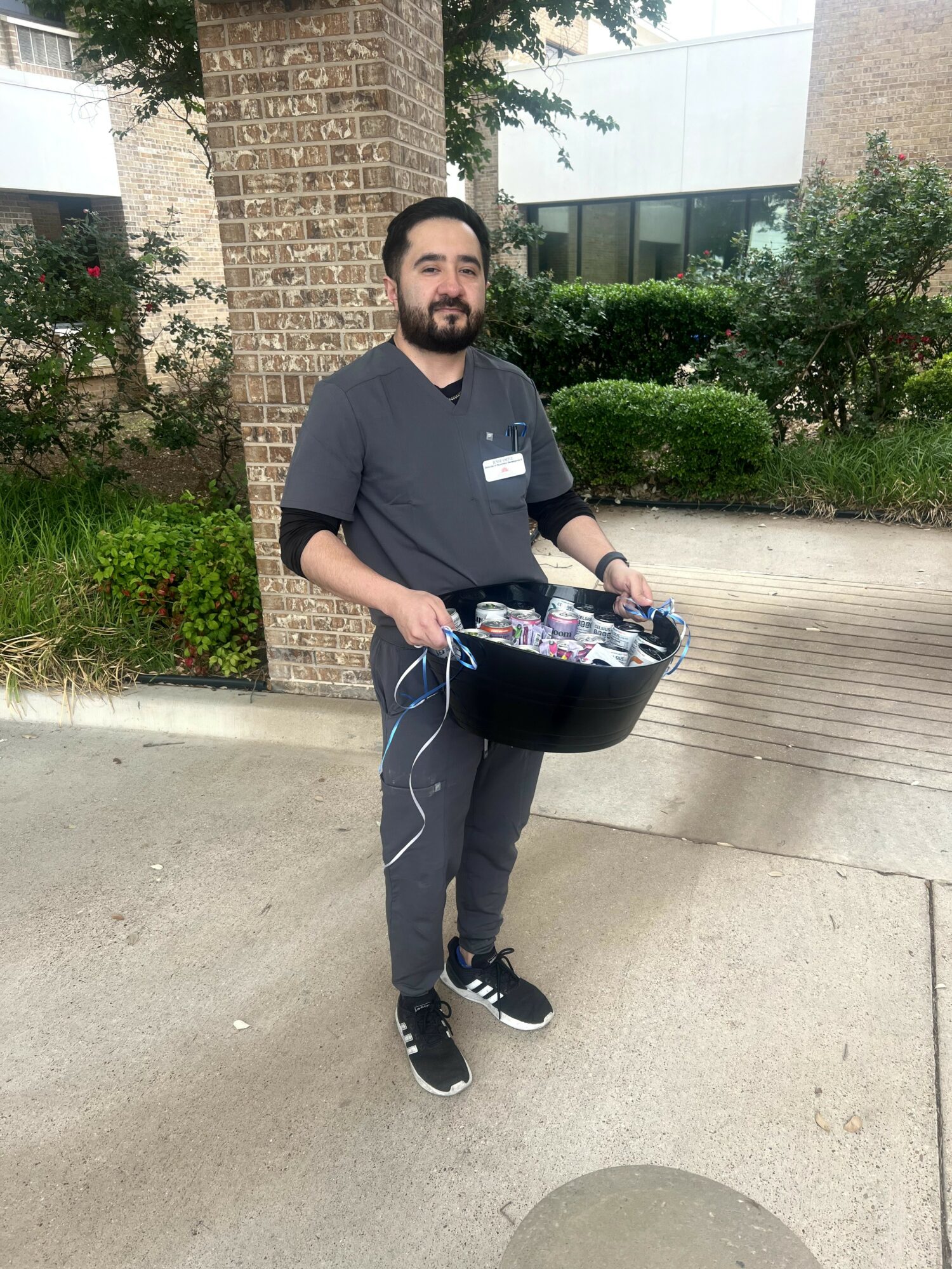 Man with beard holding a black tray of bottled drinks outdoors, standing on a sidewalk with trees and bushes behind him.