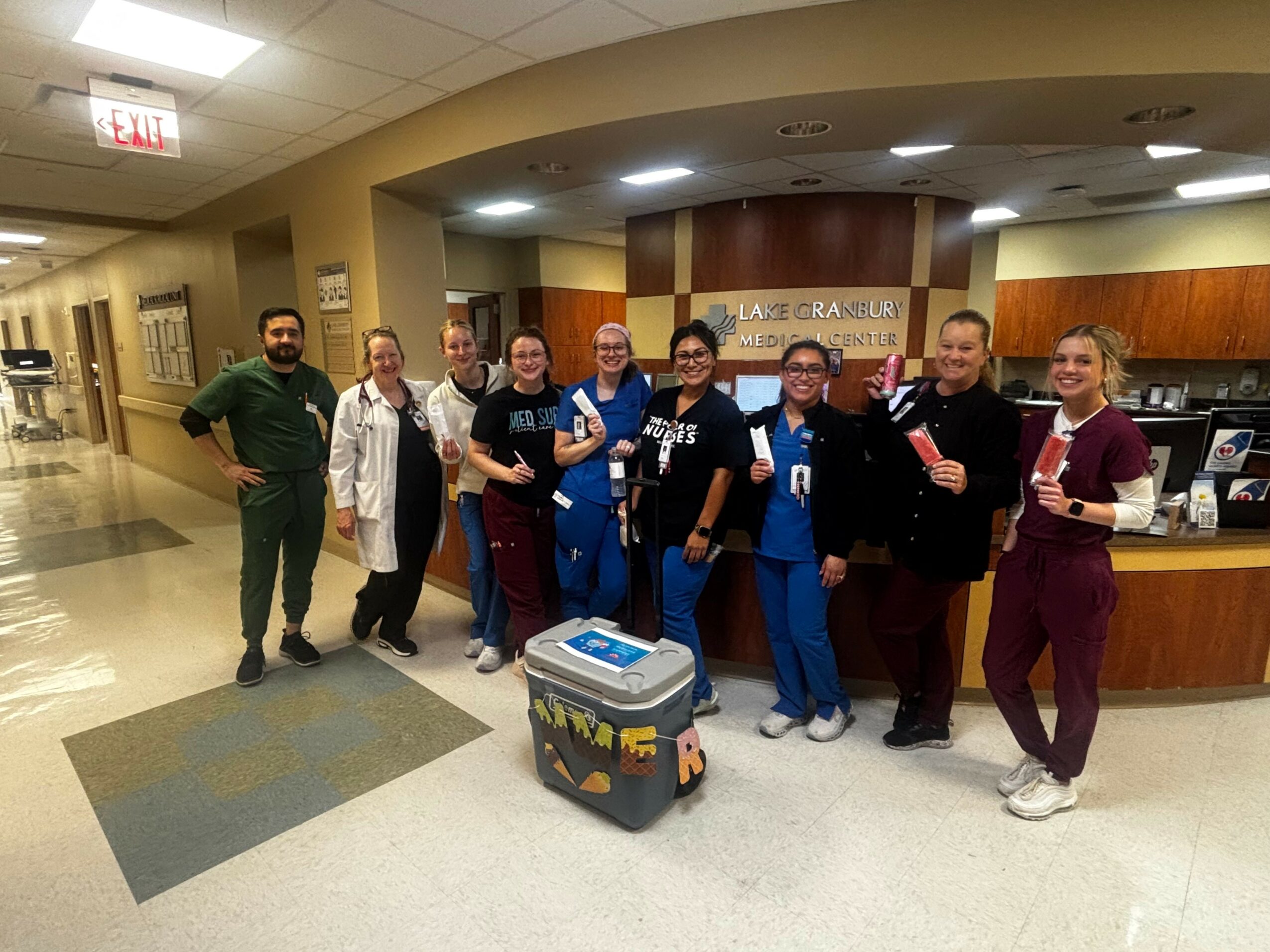Group of nine healthcare workers standing in a hospital lobby, smiling, with a cooler in front, some holding papers or folders.