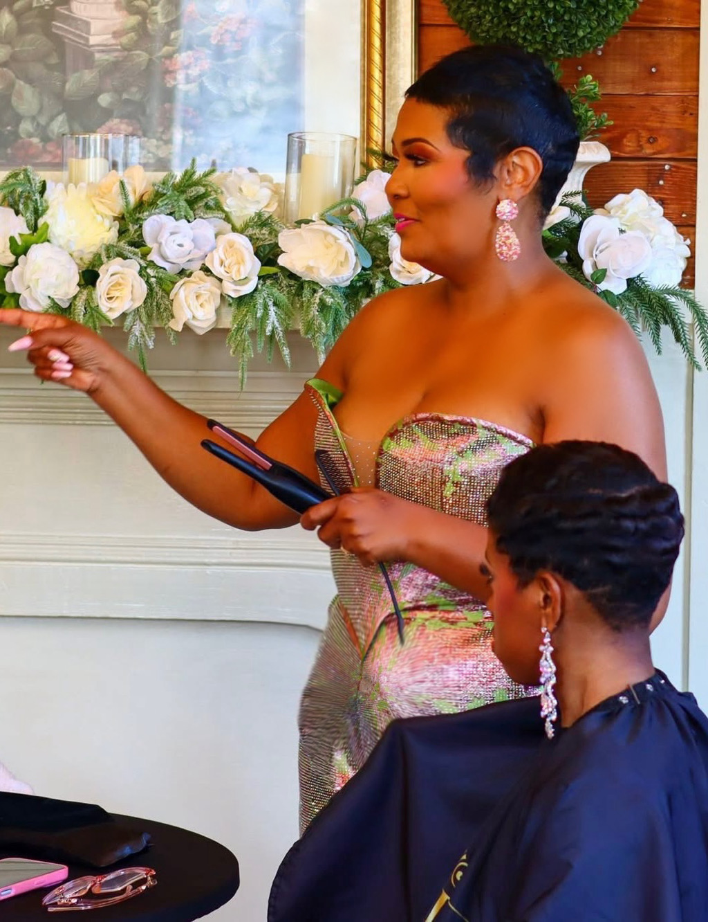 Woman in strapless dress holding a tablet, woman in dark outfit with earrings, floral arrangement in background
