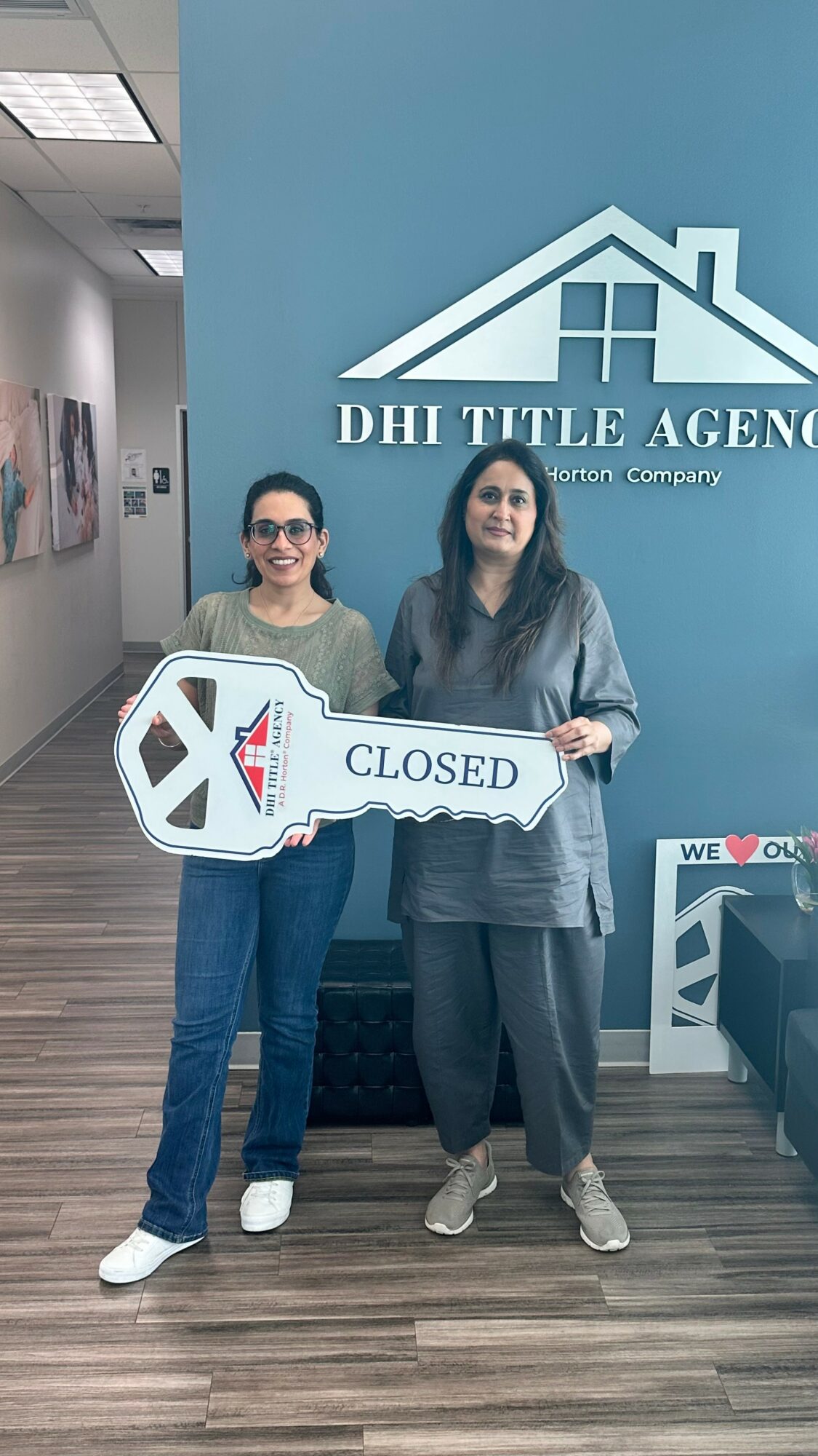 Two women standing indoors holding a large key-shaped sign that says 'CLOSED' in front of a blue wall with a house logo and text.