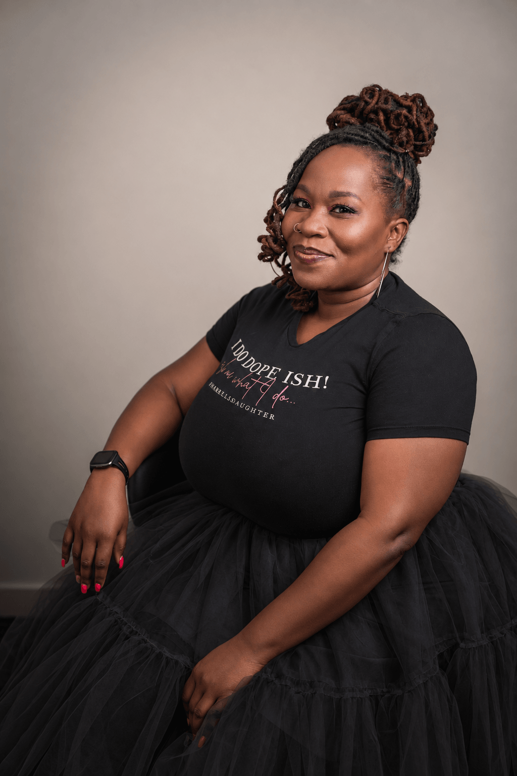 Woman with styled hair in a bun, wearing a black T-shirt and black skirt, sitting against a plain background.