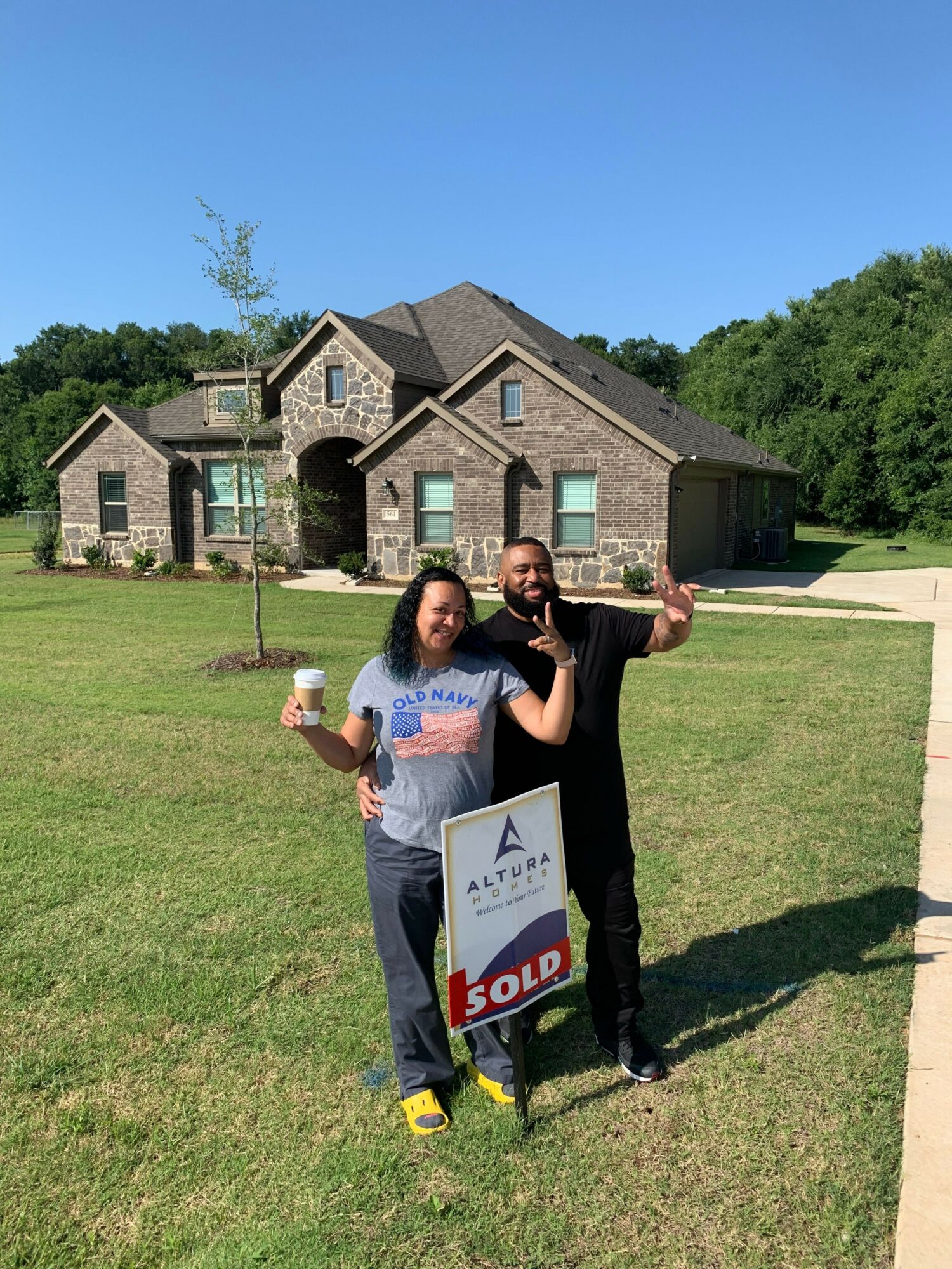 Two women standing on grass in front of a brick house, one holding a sign, the other giving a thumbs-up.