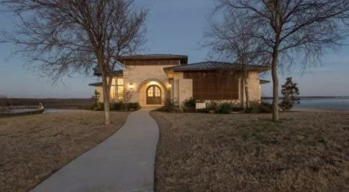 House with lit windows, two trees in front, pathway leading to entrance, near water, evening sky.