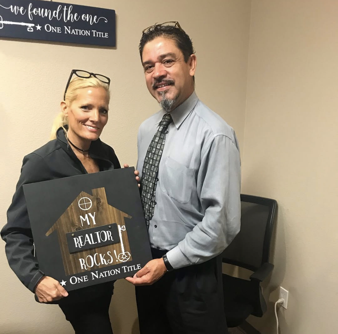 A woman and a man stand indoors, holding a sign that says 'My Realtor Rocks' with a house illustration, smiling at the camera.