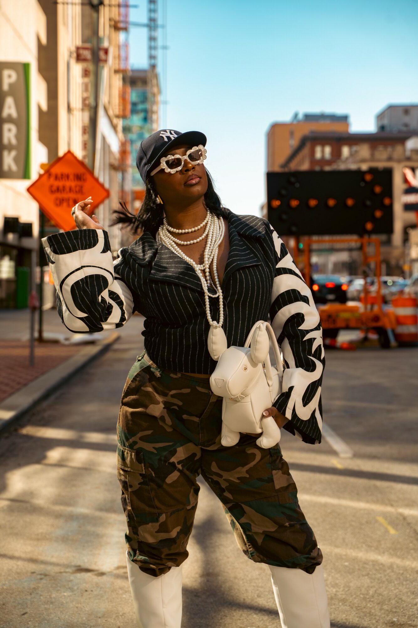 Woman in streetwear with sunglasses, jewelry, and a cap, standing on city street with buildings and traffic behind her.