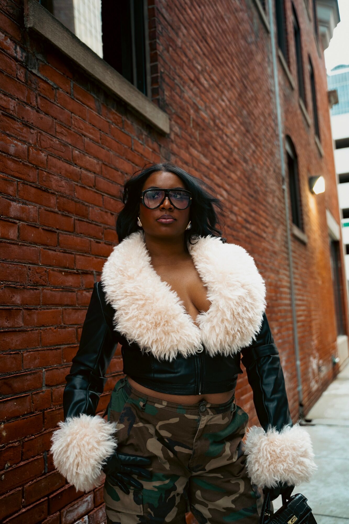 Woman with glasses and curly hair stands against a brick wall, wearing a black jacket with white fur collar and cuffs, and camouflage pants.