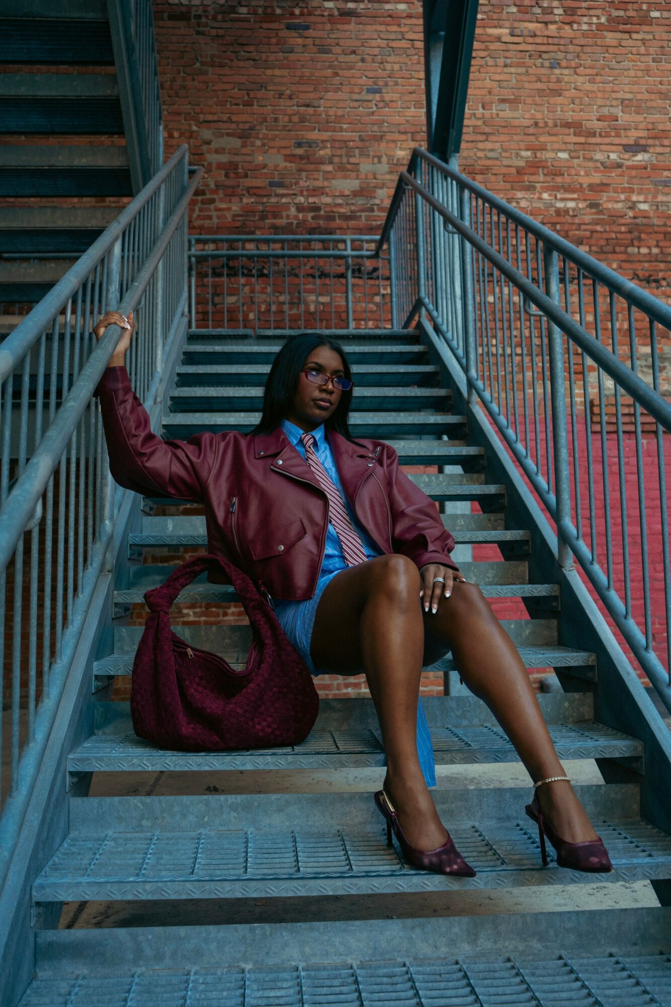 Woman sitting on outdoor metal staircase with brick wall background, holding railing, wearing a maroon jacket, blue dress, and heels.