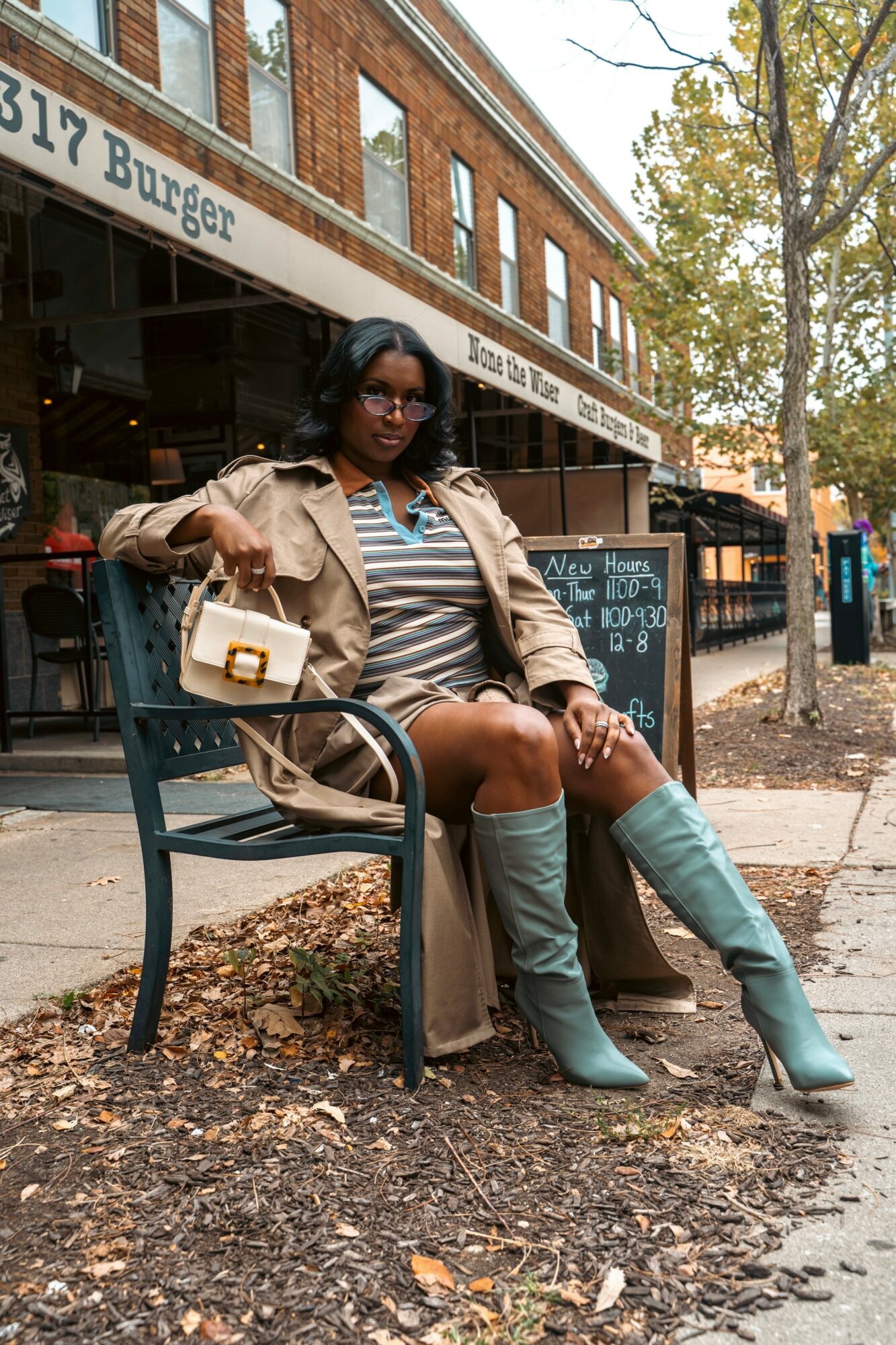 Woman sitting on a bench outside a restaurant, wearing a striped shirt, beige jacket, and knee-high boots, with trees and buildings in background.
