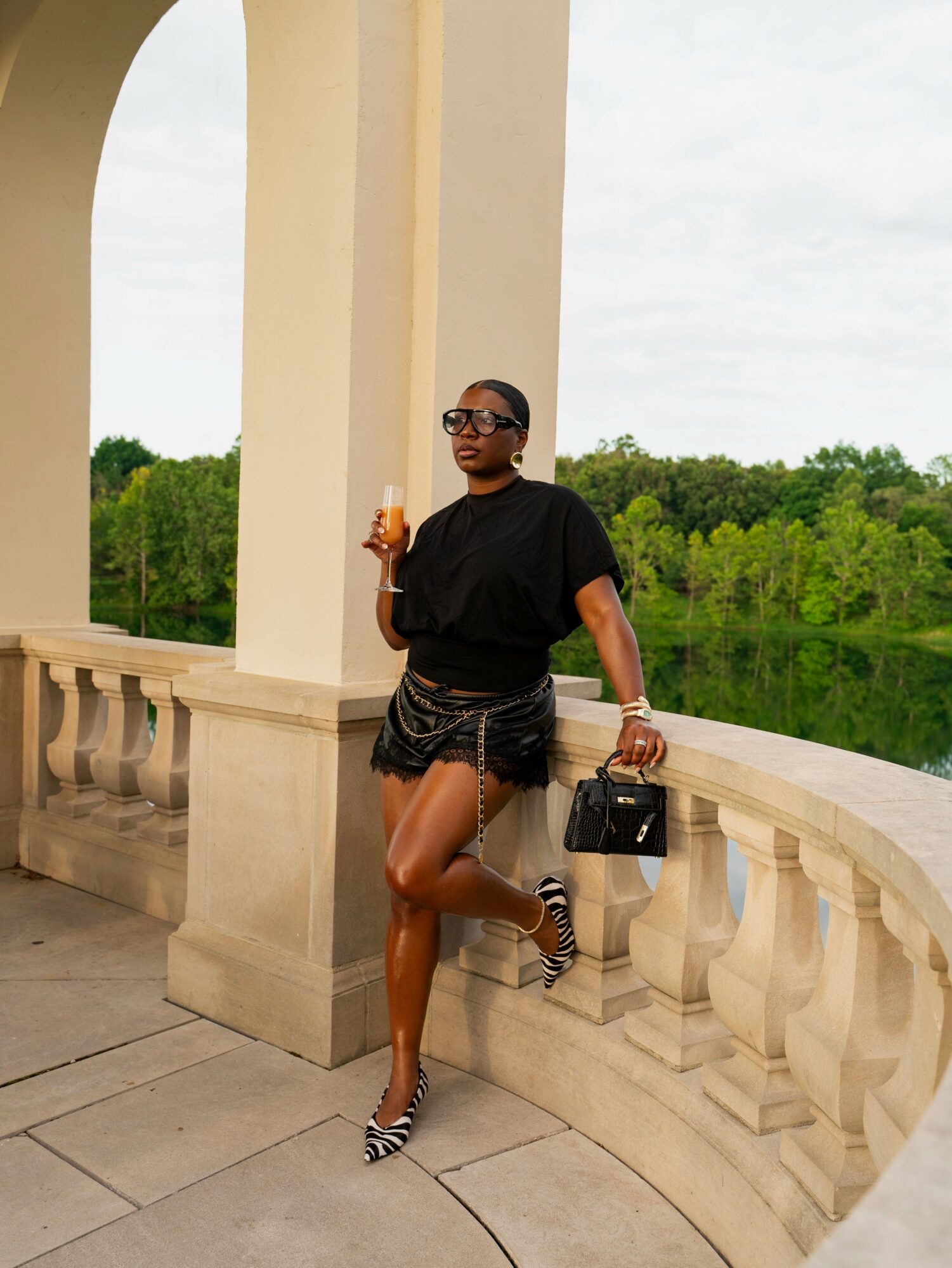 Woman in black top and patterned shorts holding a drink, standing on a balcony with a river and trees in background.