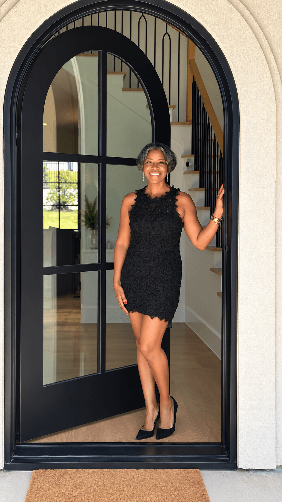 Woman in black dress standing in doorway, smiling, with staircase visible in background.