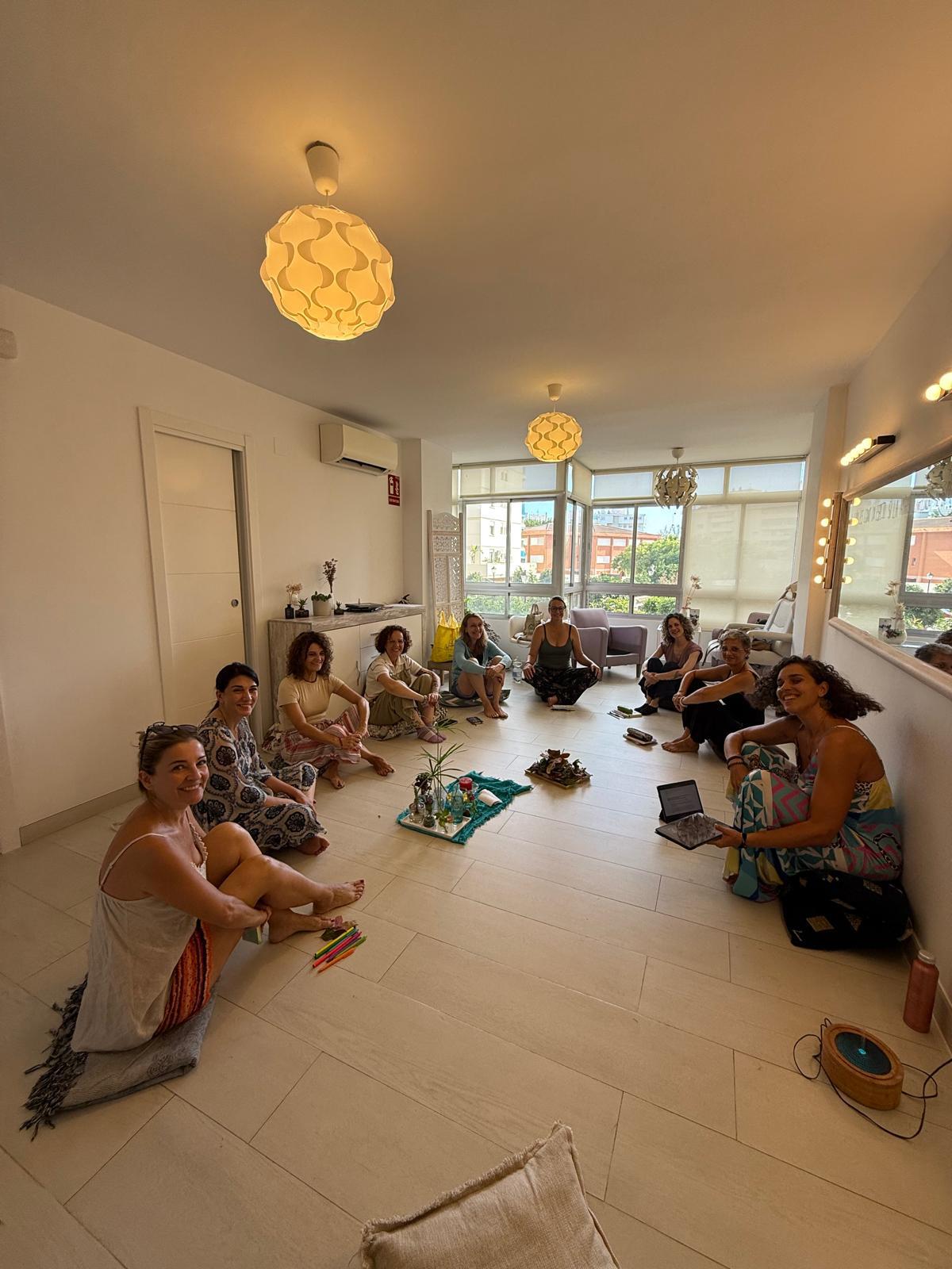 Group of people sitting on the floor in a bright room with large windows, some with laptops and bags, during daytime.