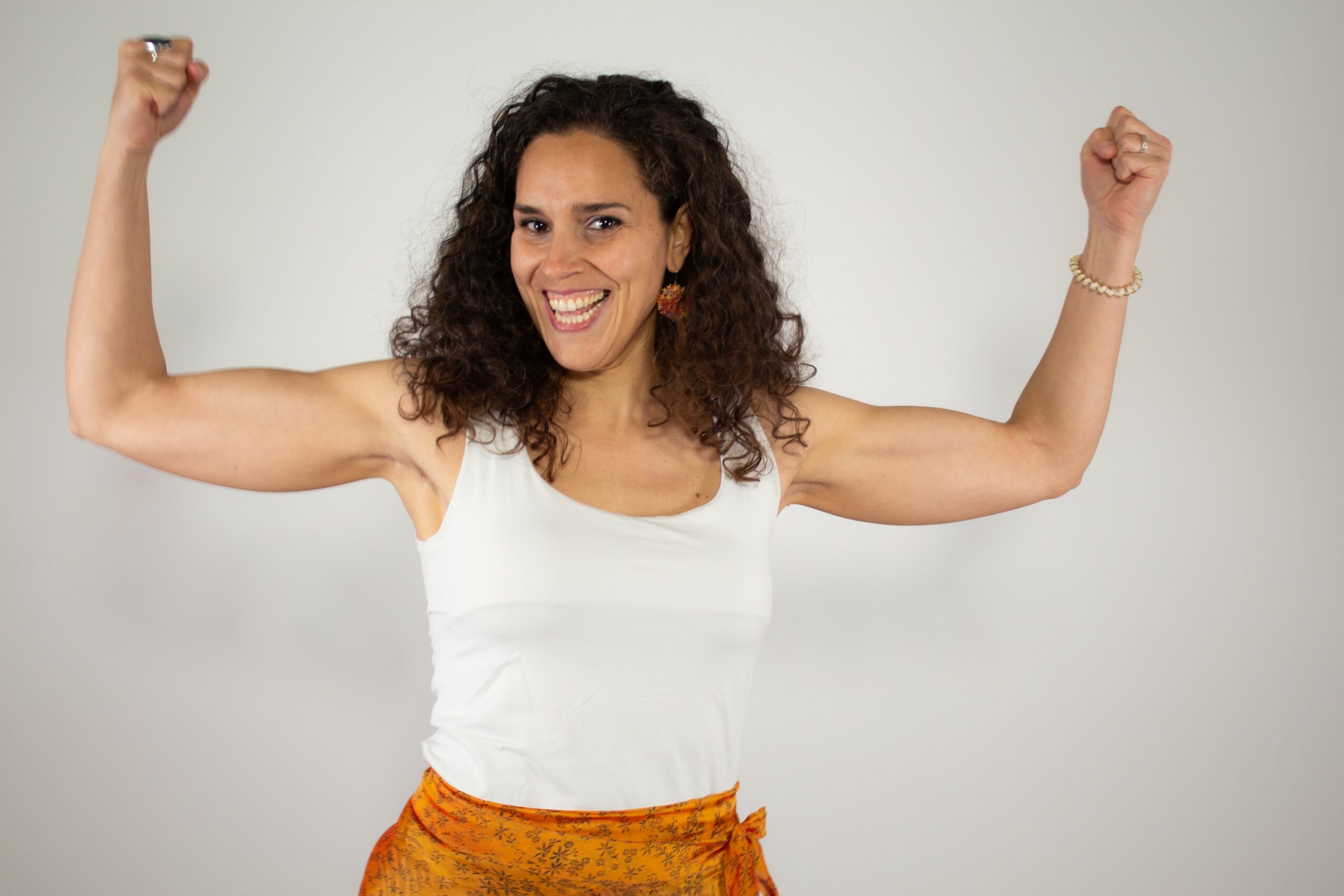 Woman with curly hair smiling and flexing her biceps, wearing a white top and orange skirt, against a plain background.