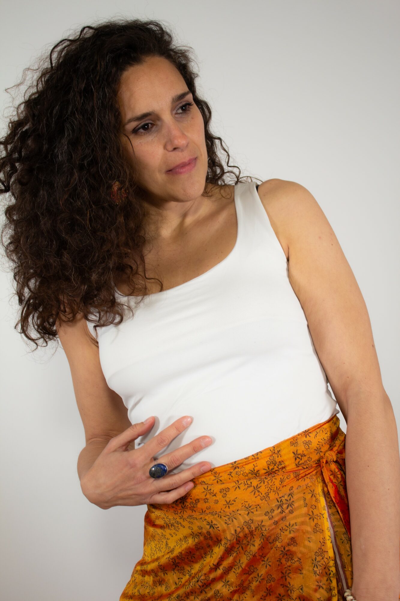 Woman with curly hair wearing a white top and orange patterned skirt, looking to the side, holding her stomach.