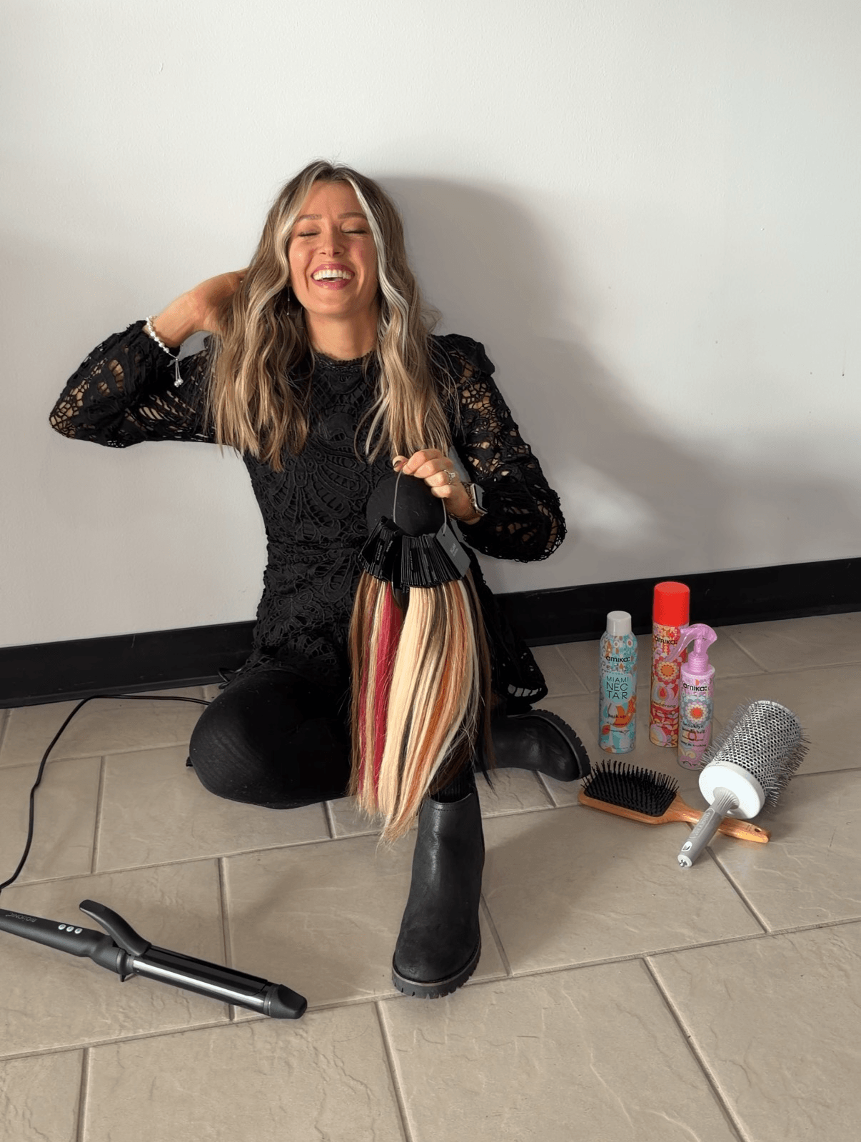 Woman sitting on the floor, smiling, holding a striped bag, with beauty products and a hairdryer nearby, against a plain wall.