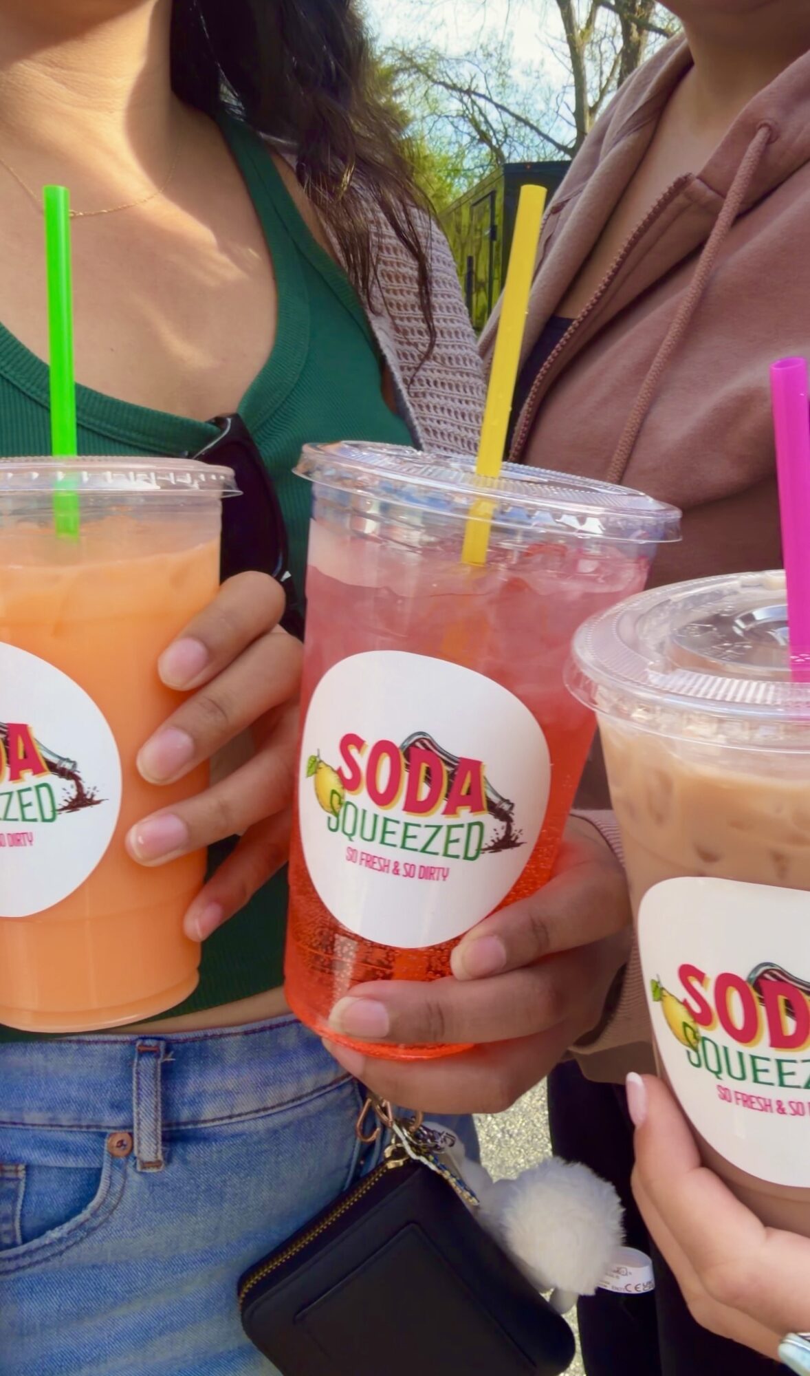 Three people holding colorful drinks with straws, close-up of their hands and torsos outdoors.