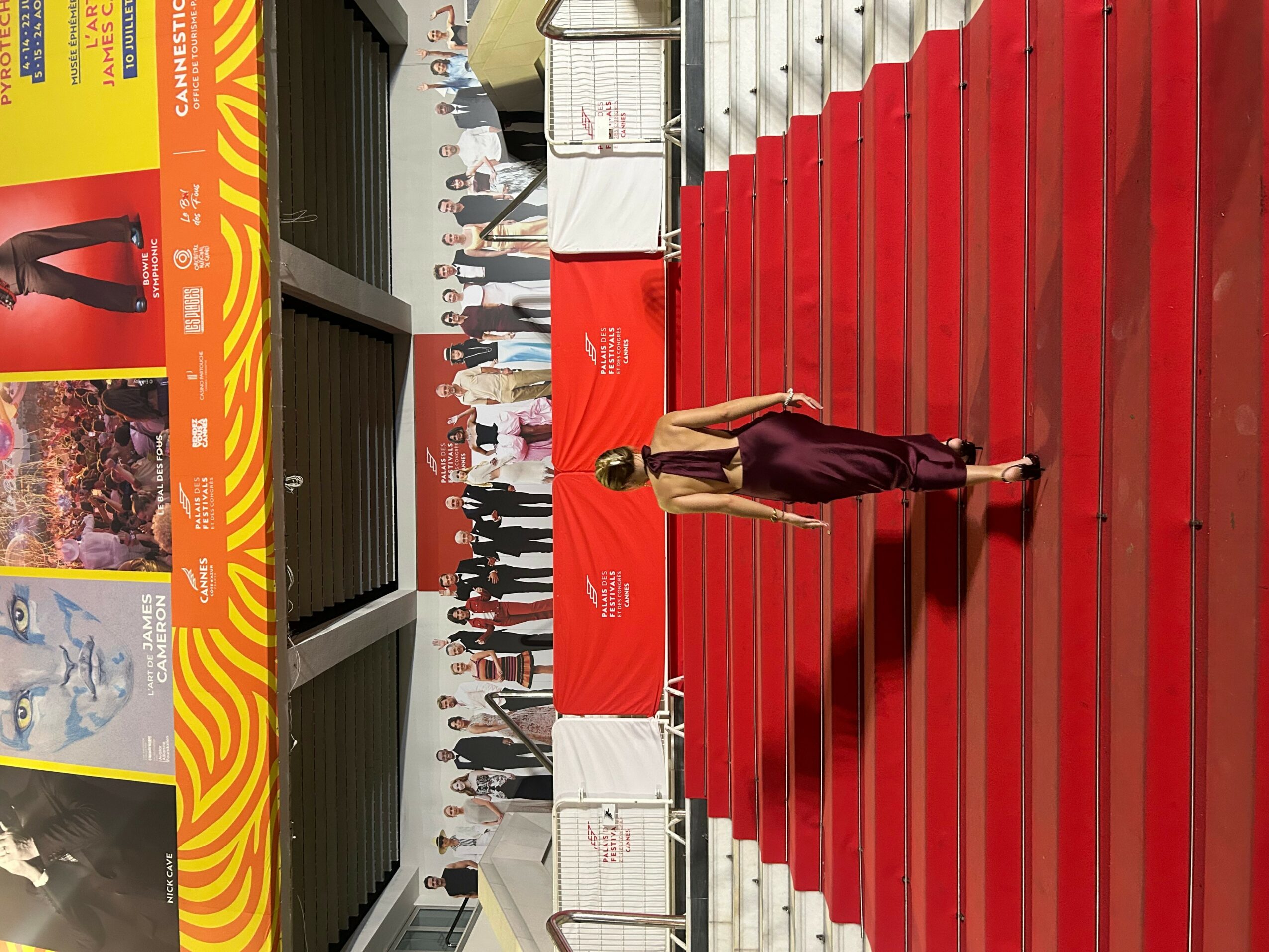 Person walking on red carpet at event with crowd and banners in background.