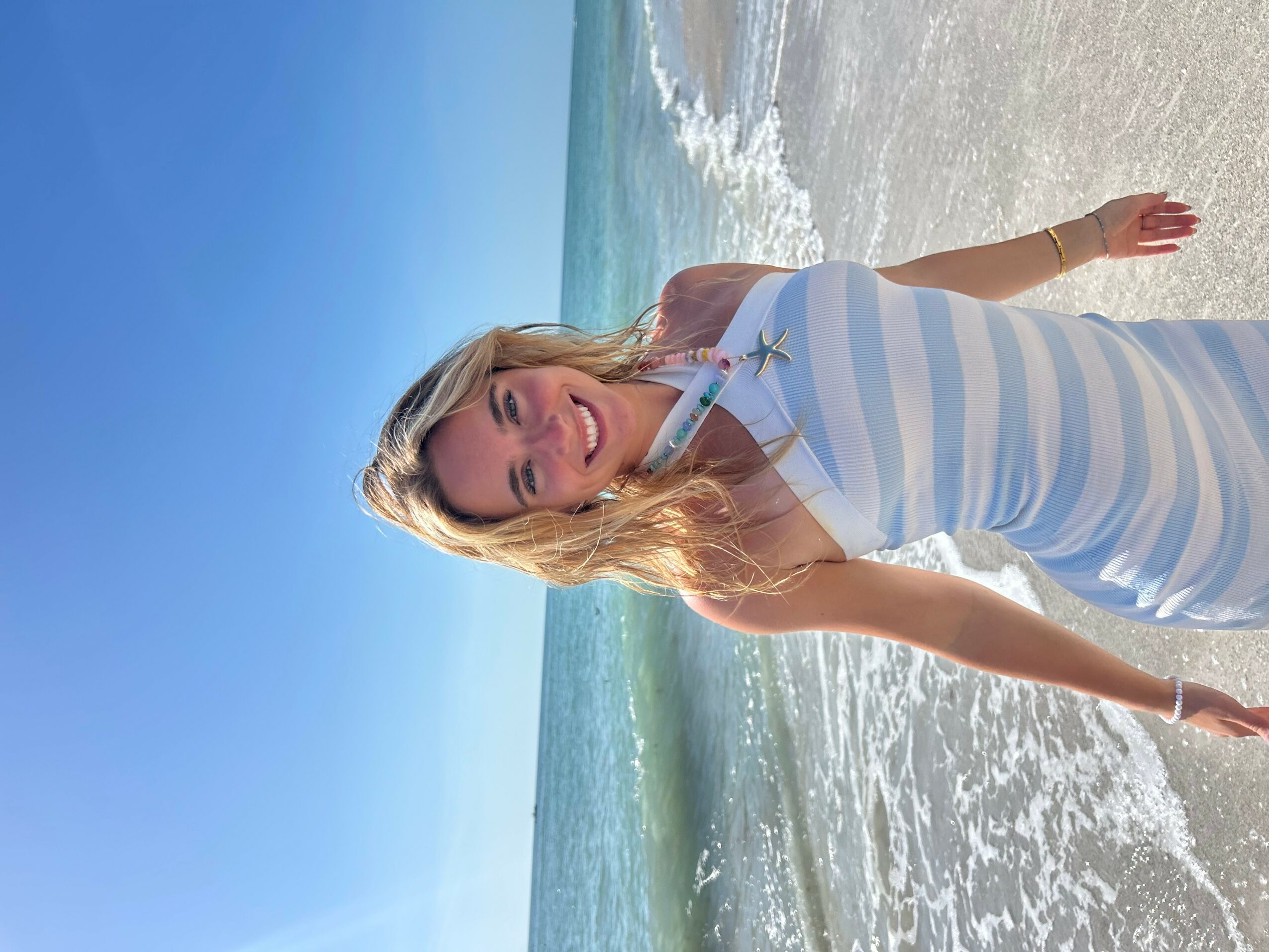 Woman in a striped dress standing in shallow ocean water at the beach, smiling, with blue sky and ocean in background.