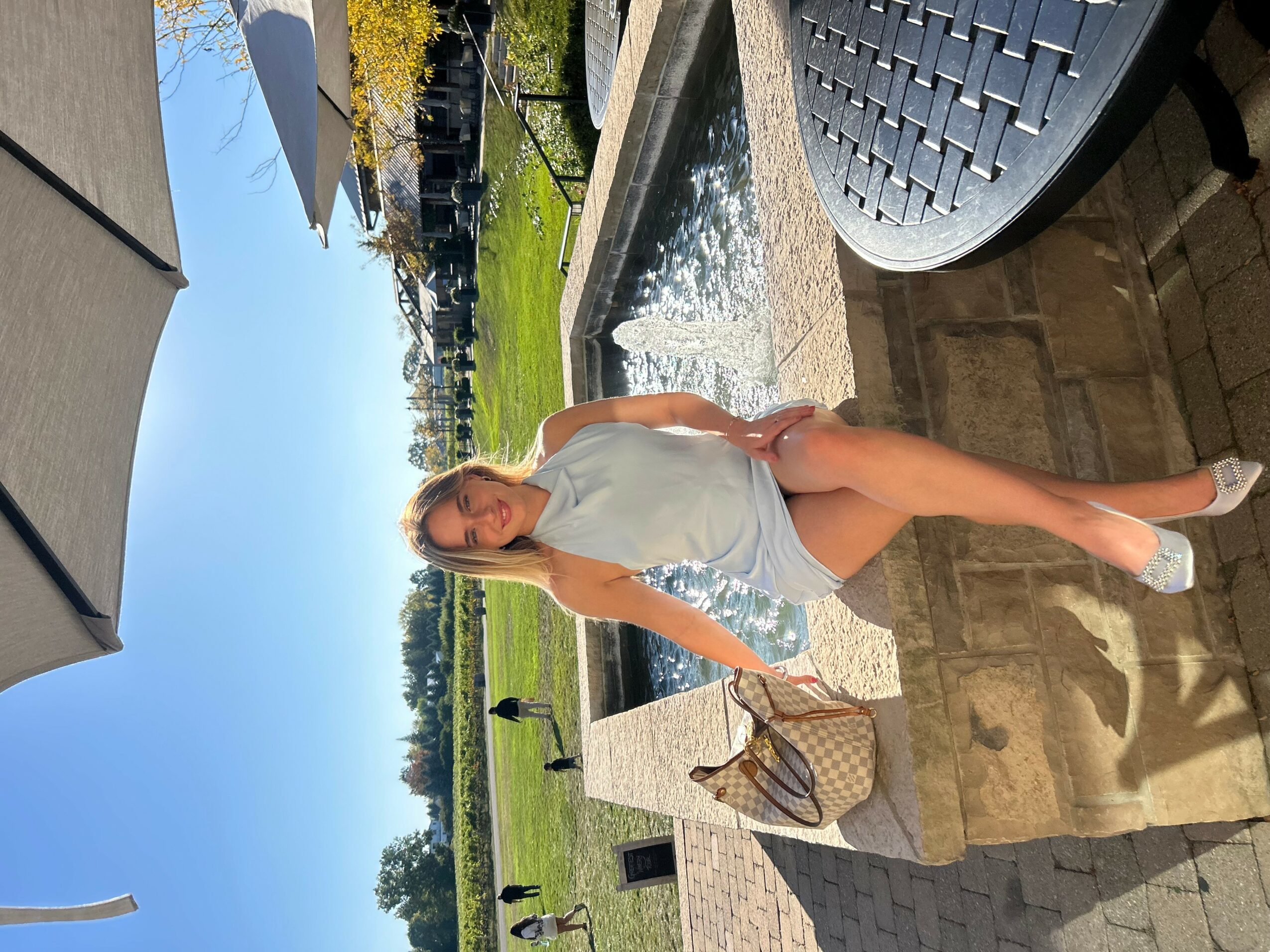 Woman in white dress standing outdoors near a fountain, holding a bag, with a garden and blue sky in background.