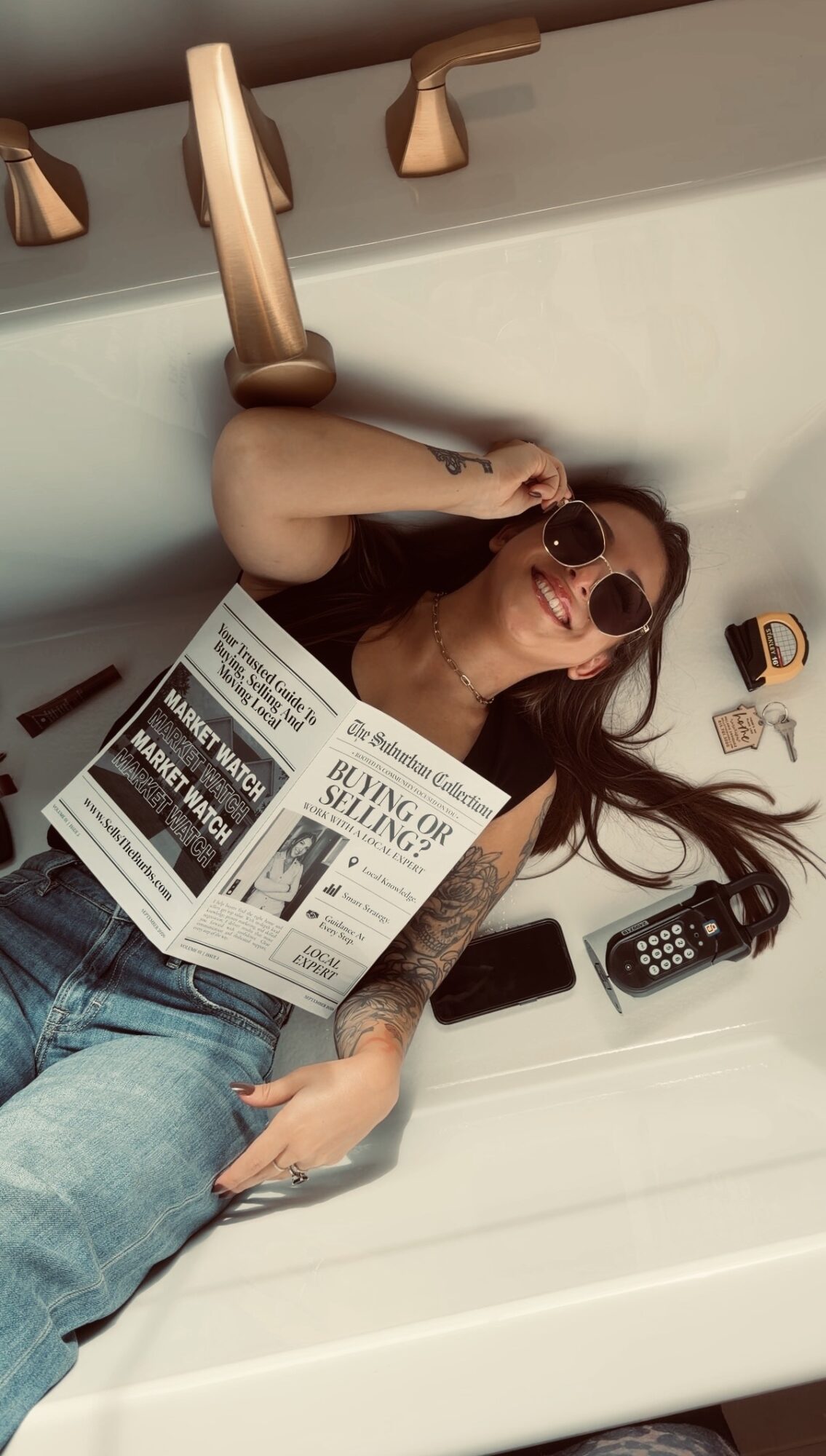 Person with sunglasses lying in a bathtub, holding a newspaper, with a remote control and other items nearby.