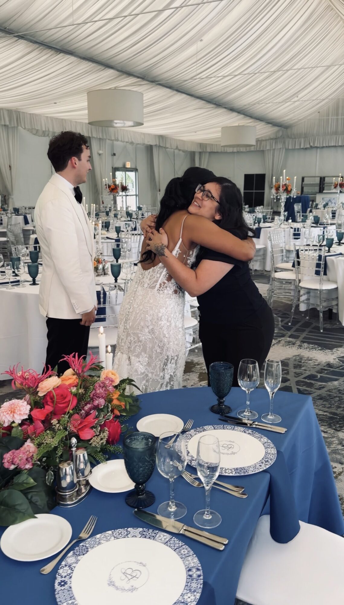 Two women hug at a formal event, with a man in a white suit observing nearby, decorated table in foreground.