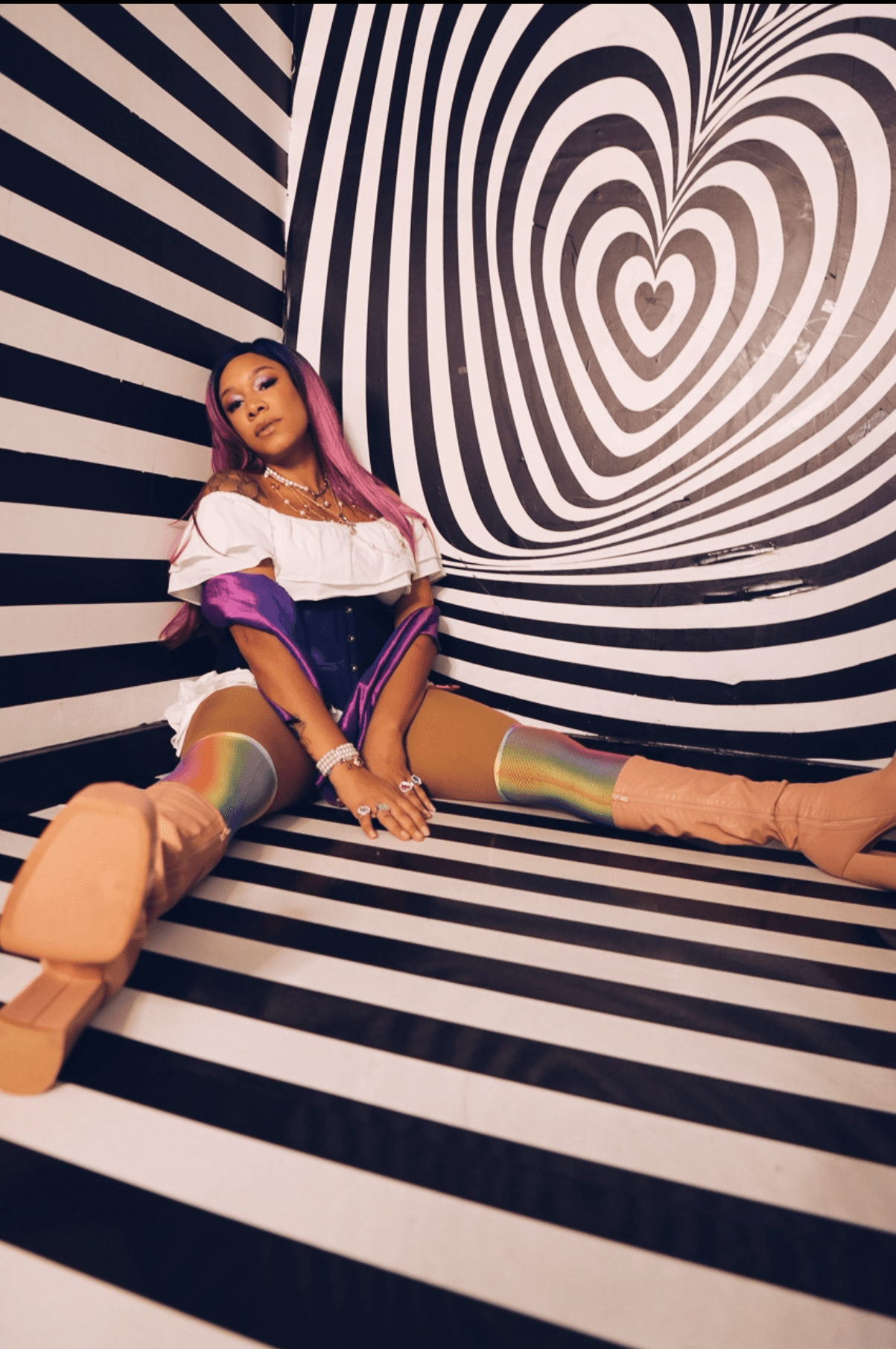 Young woman with pink hair sitting on black and white striped floor and wall with heart-shaped pattern, wearing colorful clothing and boots.