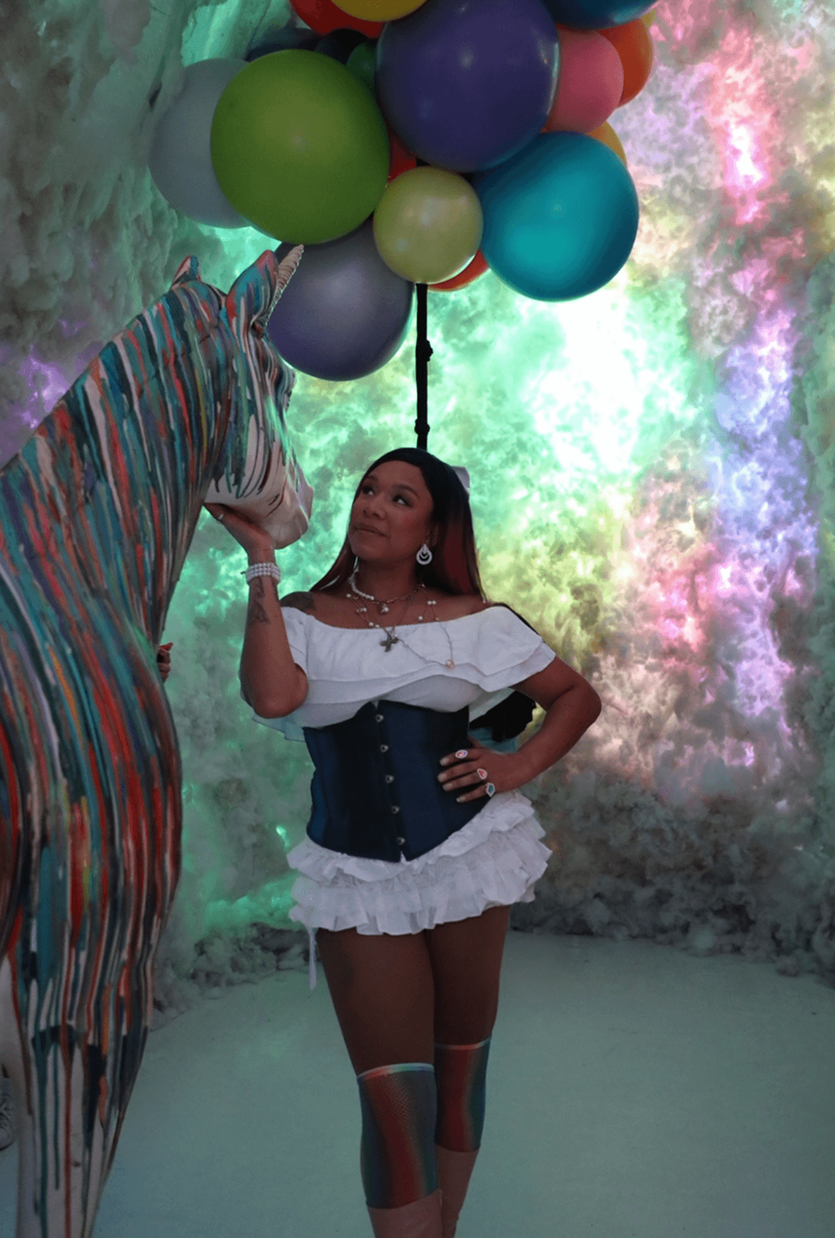 Person holding colorful balloons with a girl in a white top, black corset, and ruffled skirt standing in front of a vibrant background.