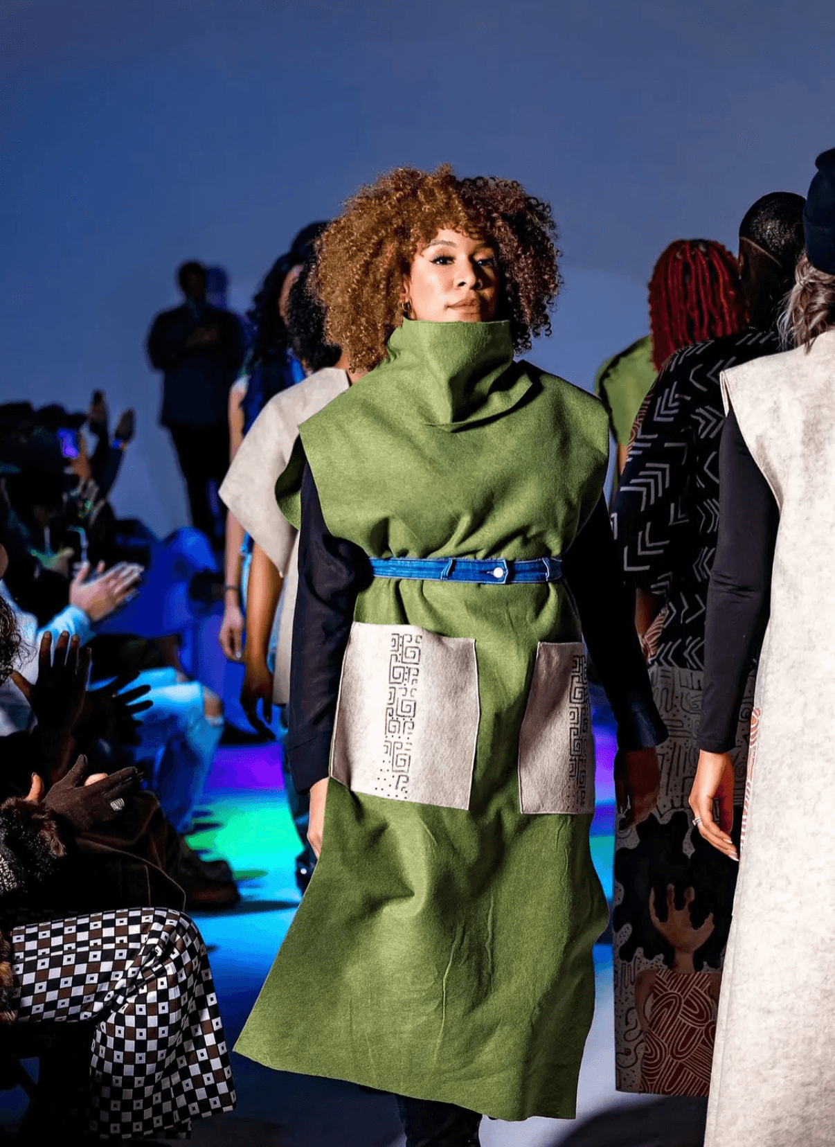 Model walking on runway wearing a green dress with large collar and blue belt, audience seated on sides, evening setting.