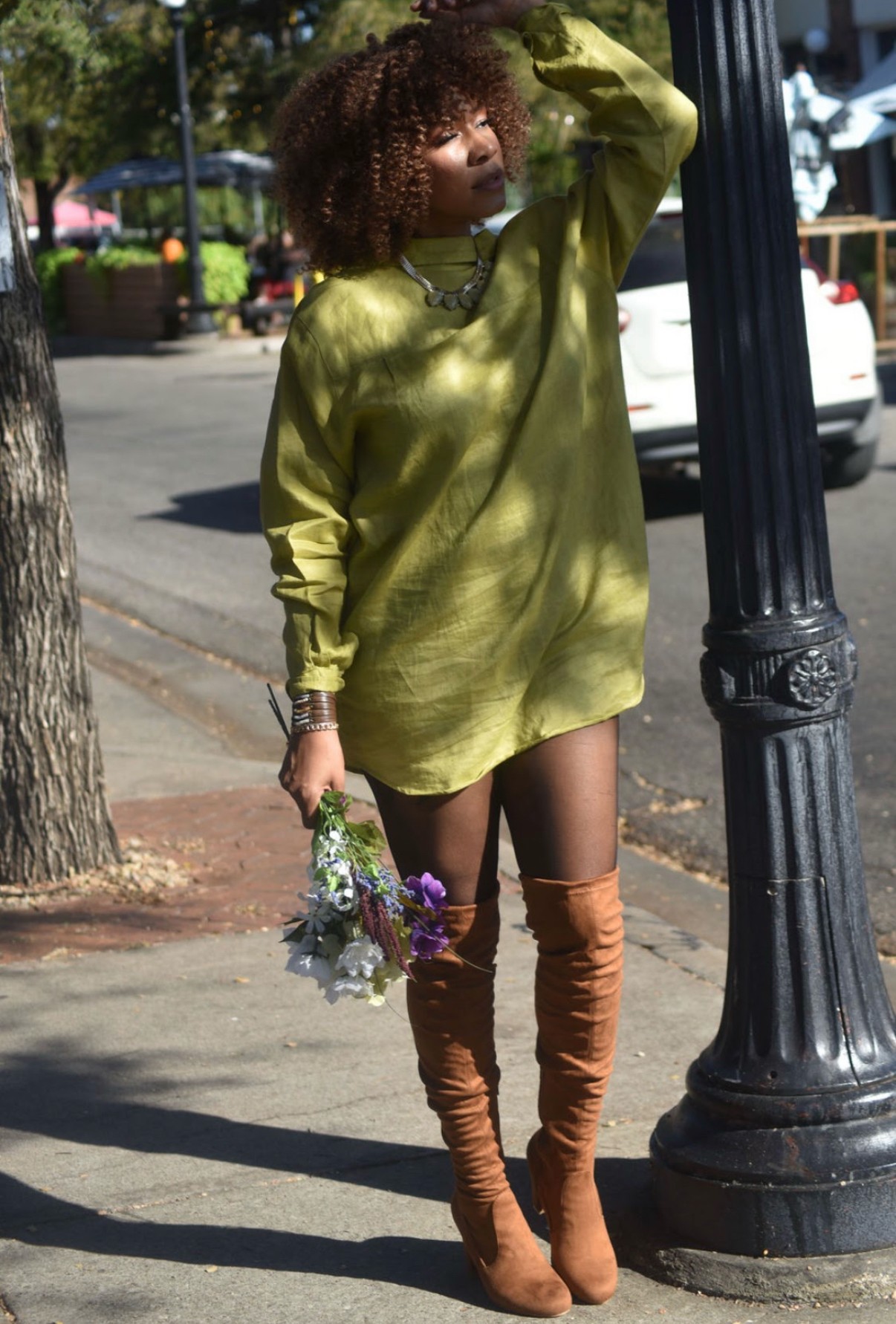 Person with curly hair in an oversized green sweater dress, brown thigh-high boots, holding a bouquet, standing outdoors near a lamppost.