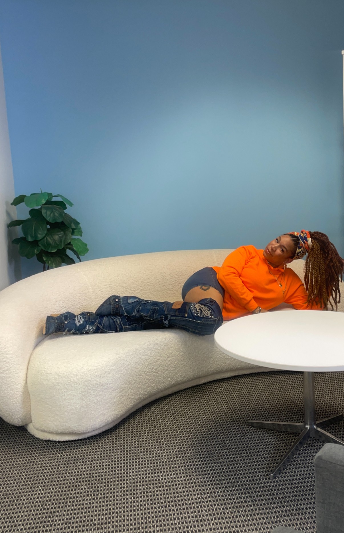 Person lying sideways on a cream sofa, wearing an orange top and ripped jeans, with a blue wall and plant in background.