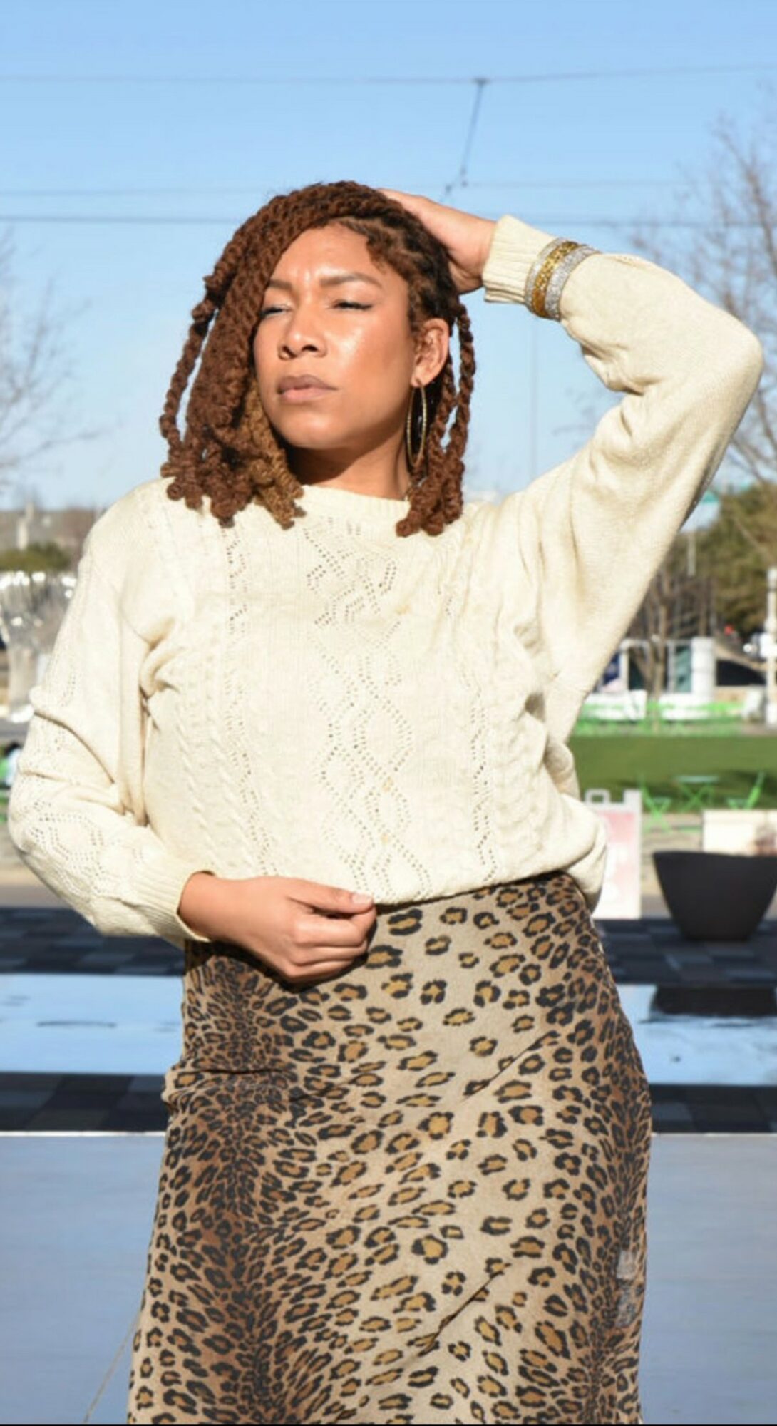 Woman with curly hair wearing a cream sweater and leopard print skirt, standing outdoors with her hand on her head.