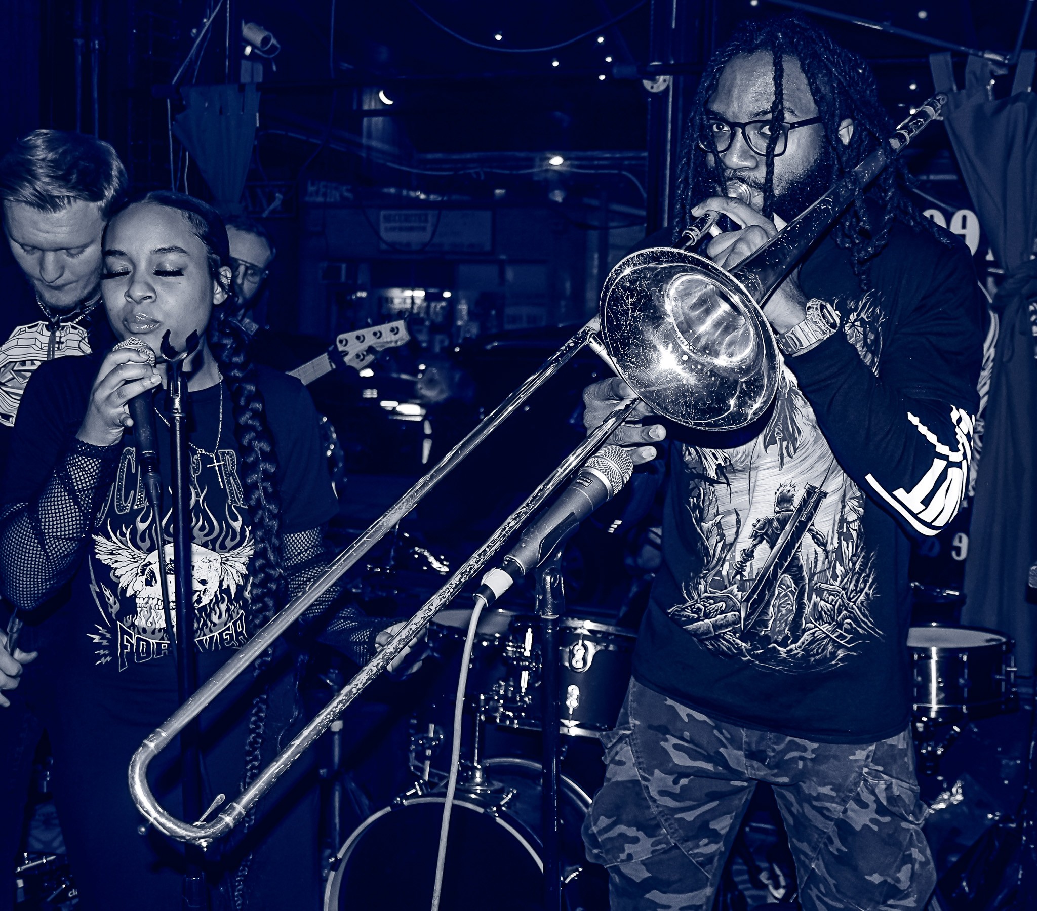 Two children playing musical instruments, one with a microphone and the other with a trombone, in a dimly lit setting.