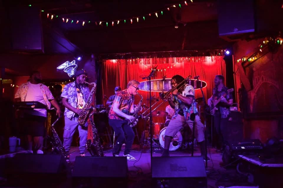 Group of musicians performing on stage with red curtain backdrop, colorful lights, and string lights overhead.