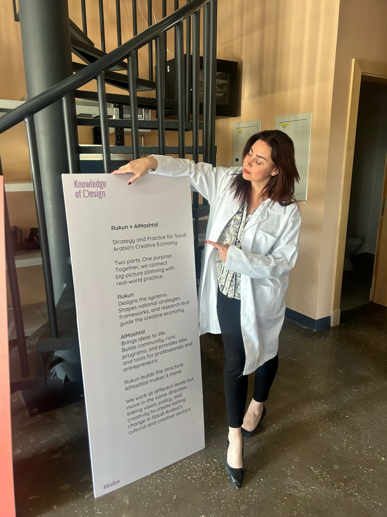 Woman with shoulder-length hair in a white coat, standing next to a large informational sign, smiling and pointing at it.