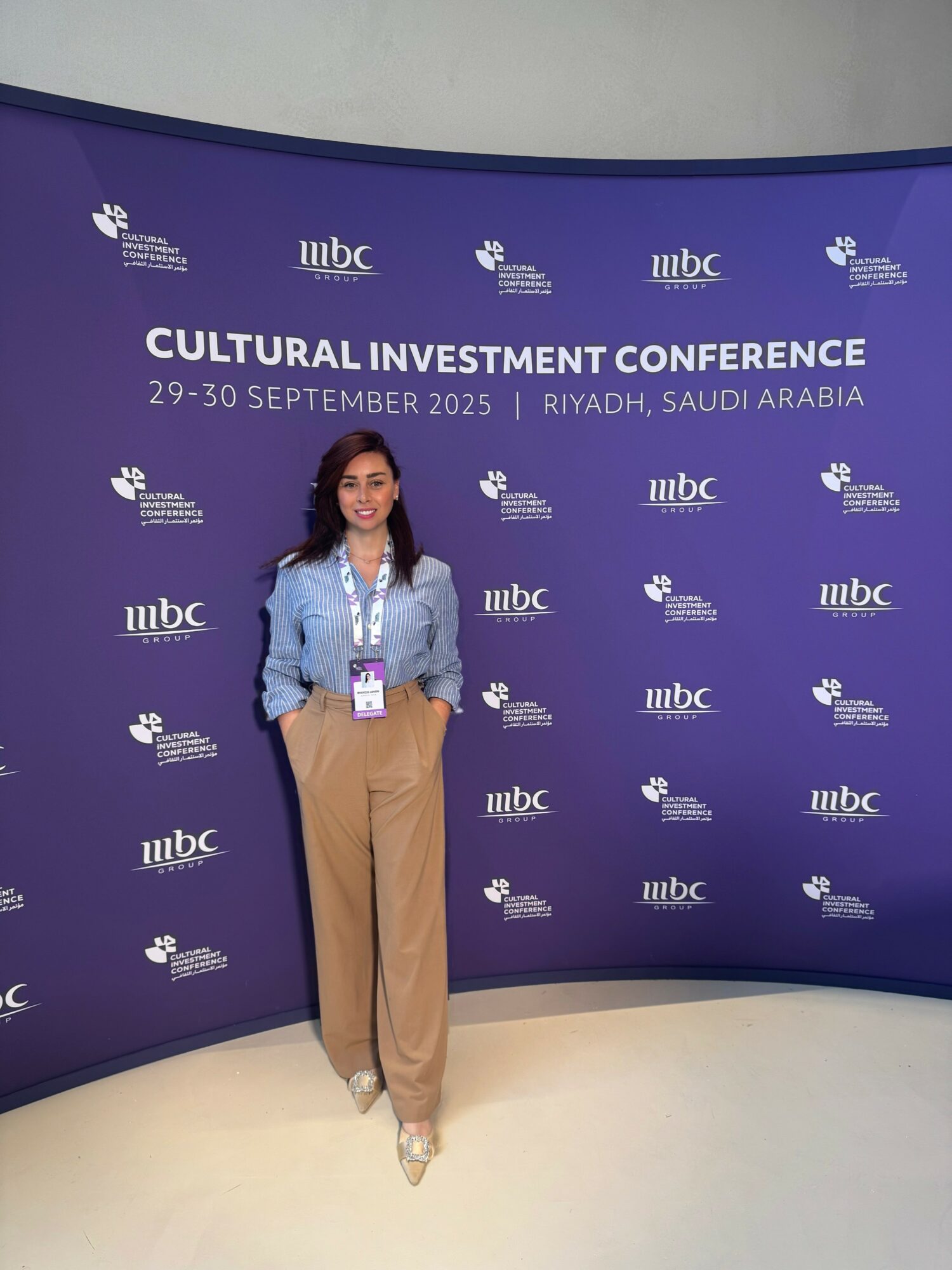 Woman standing in front of a purple backdrop at the Cultural Investment Conference in Riyadh, Saudi Arabia, September 2025, smiling, wearing beige pants and a blue shirt.
