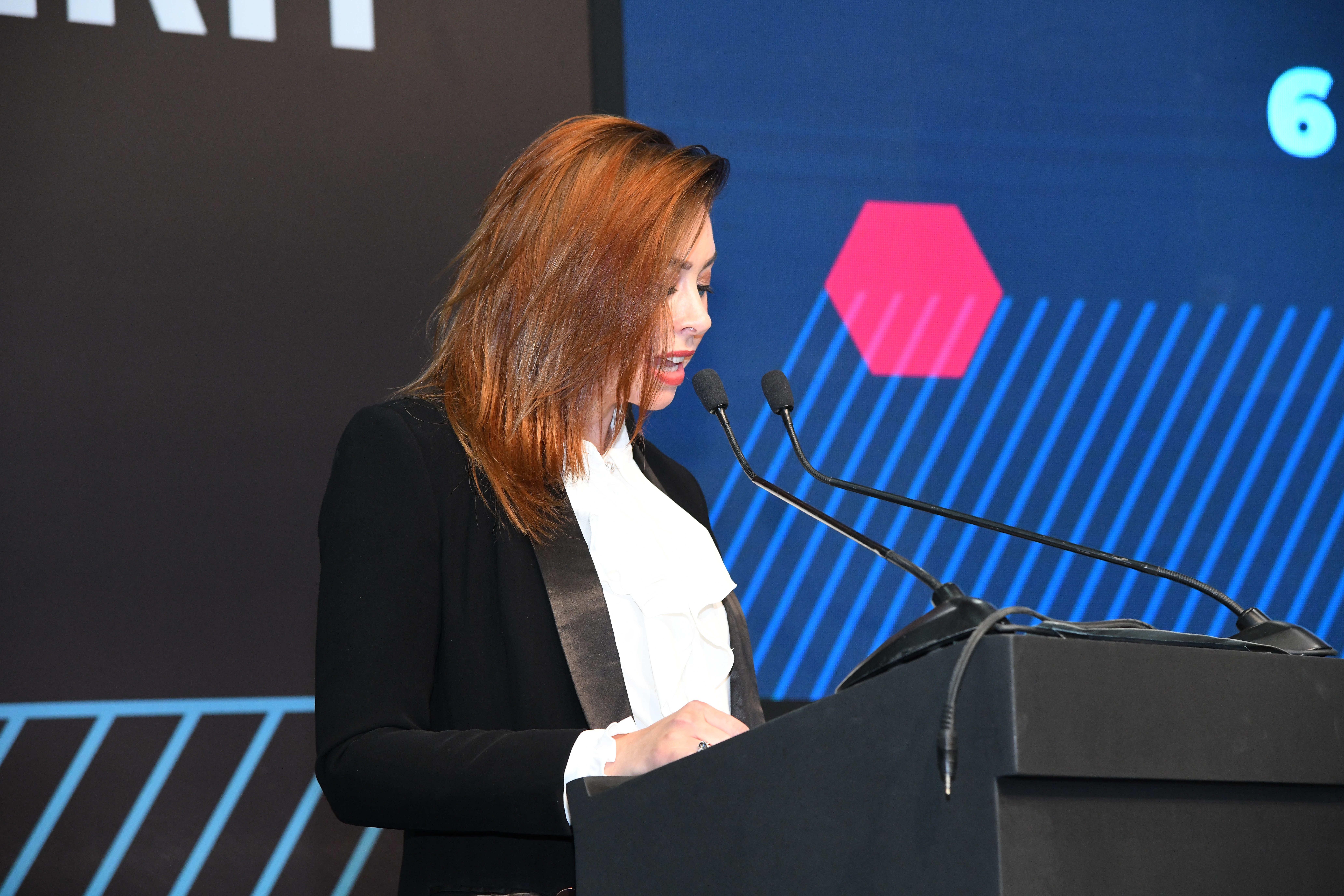 Woman with red hair speaking at a podium with a blue background and geometric shapes
