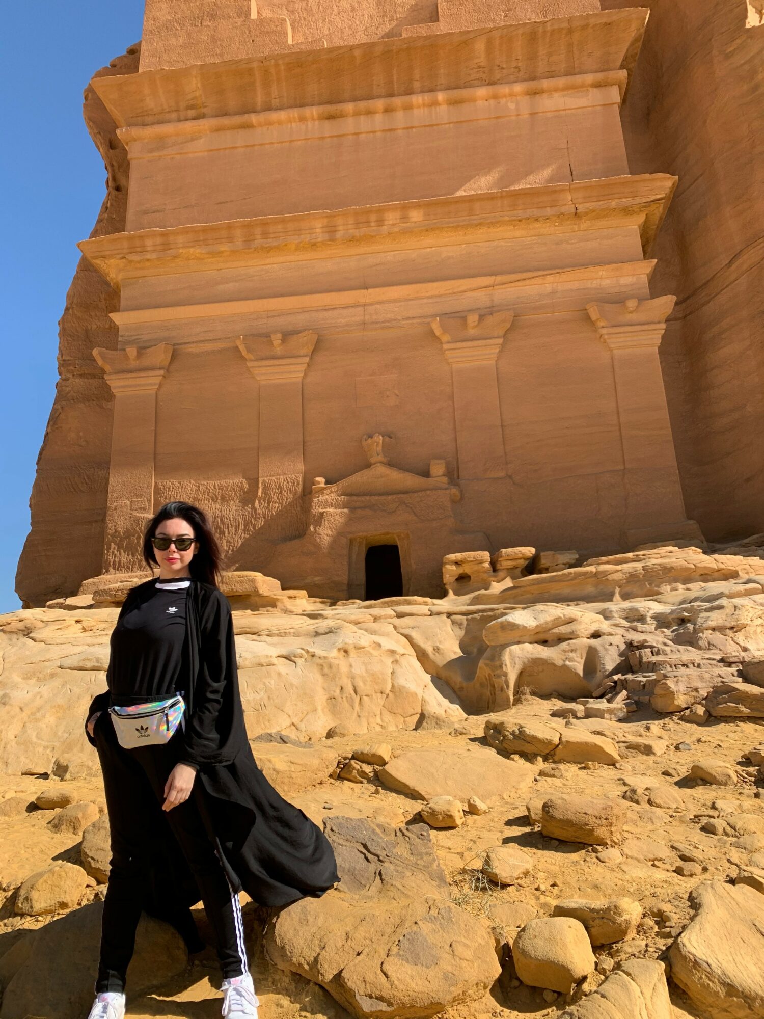 Person wearing black clothing and sunglasses standing on rocky ground in front of a large ancient stone structure.