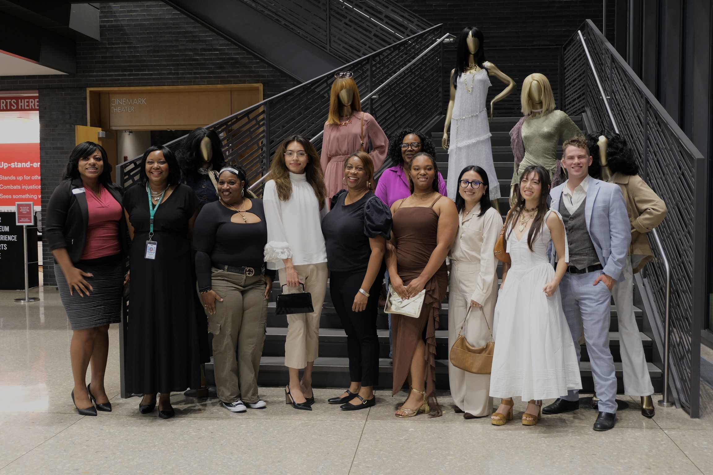 Group of people standing together indoors, some holding bags, with stairs and a railing in the background.