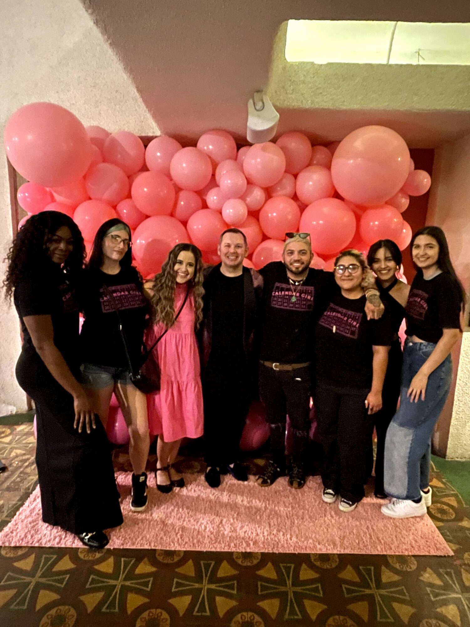 Group of seven people standing in front of pink balloons and a wall, smiling, indoors.