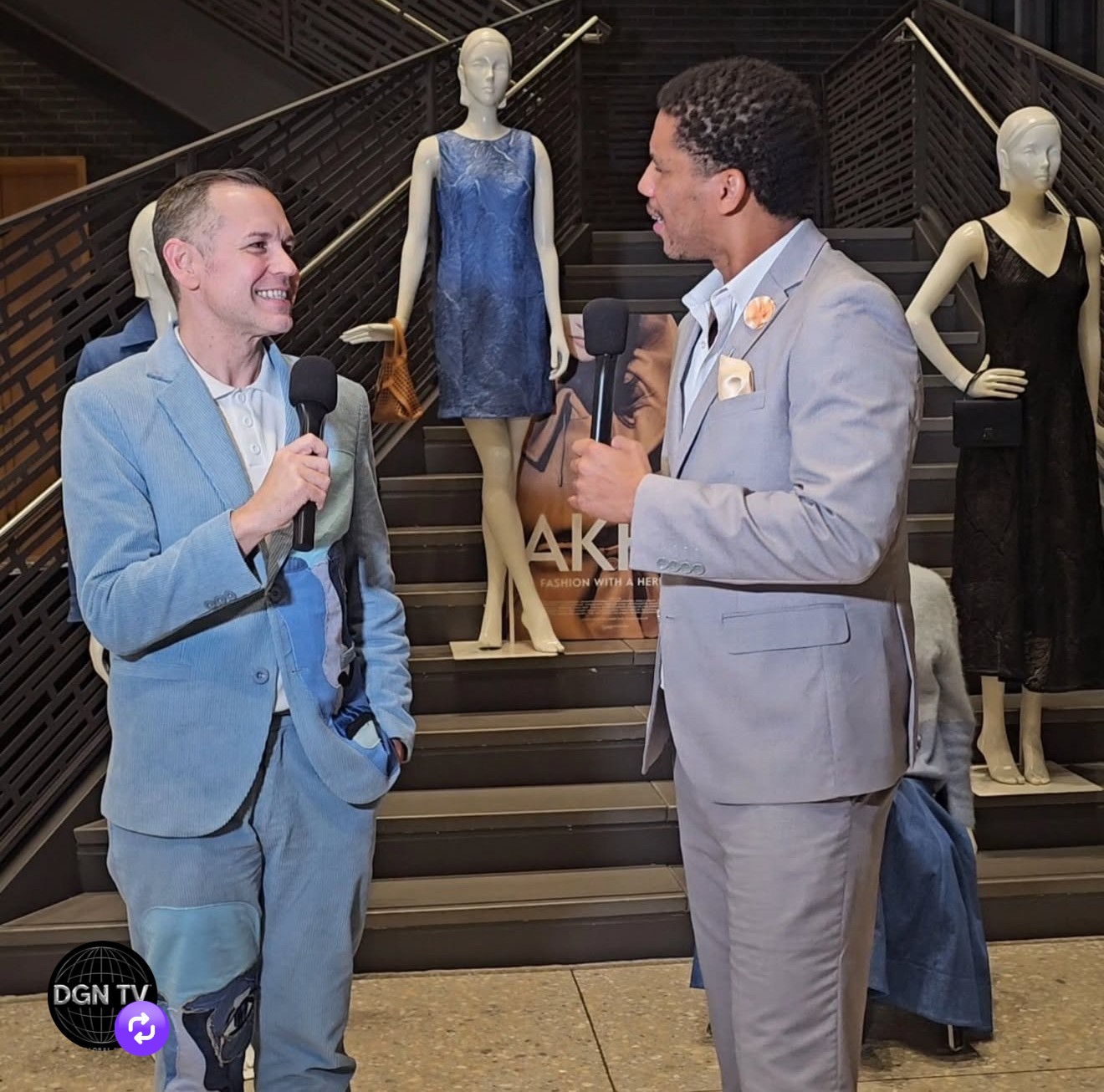Two men in suits holding microphones and talking, mannequins with dresses in the background, staircase behind them.