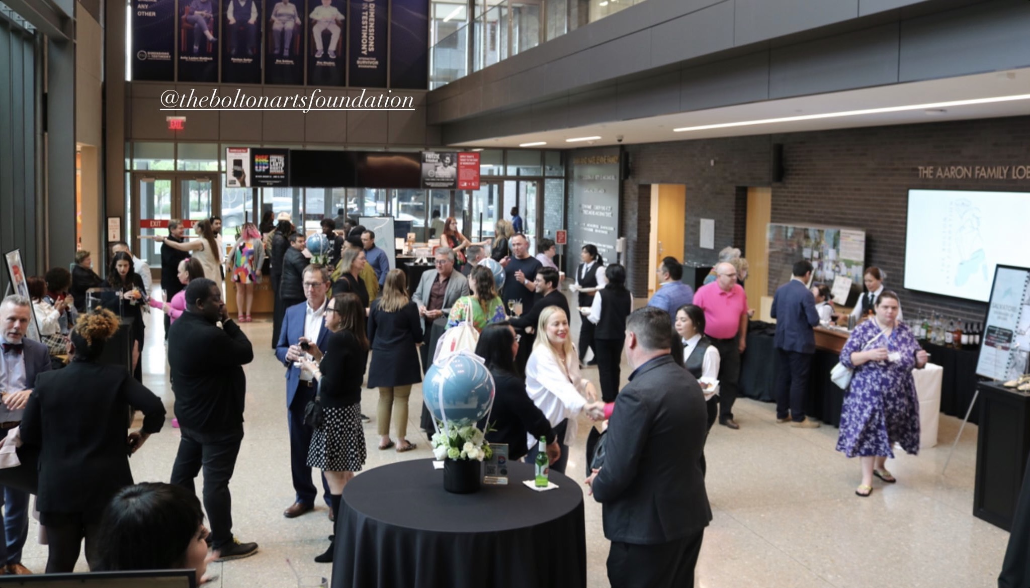 Crowd of people in a spacious indoor area with large windows, some talking, some walking, and a table with a globe.