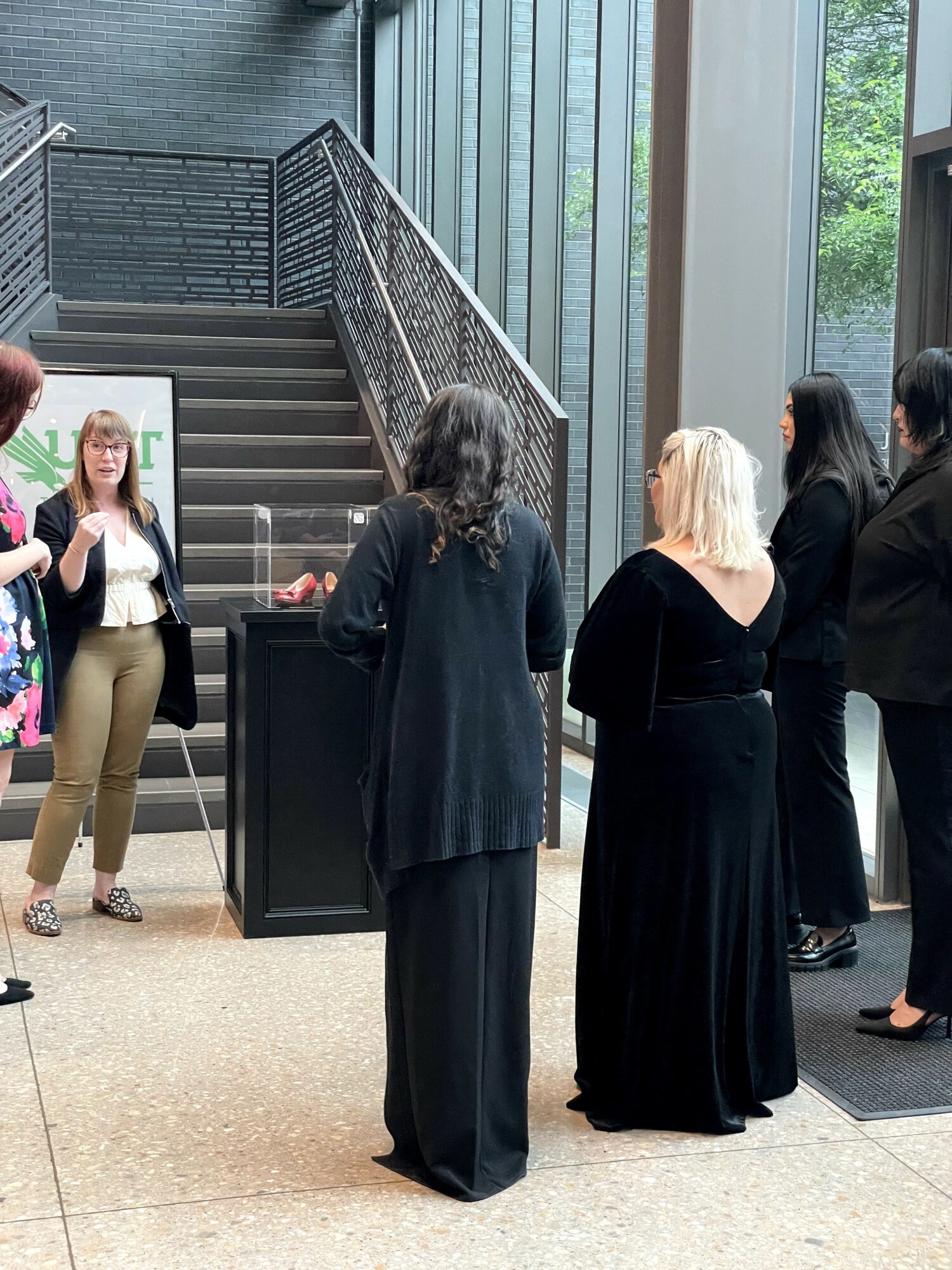 Group of people standing indoors near stairs and large windows, engaged in conversation.