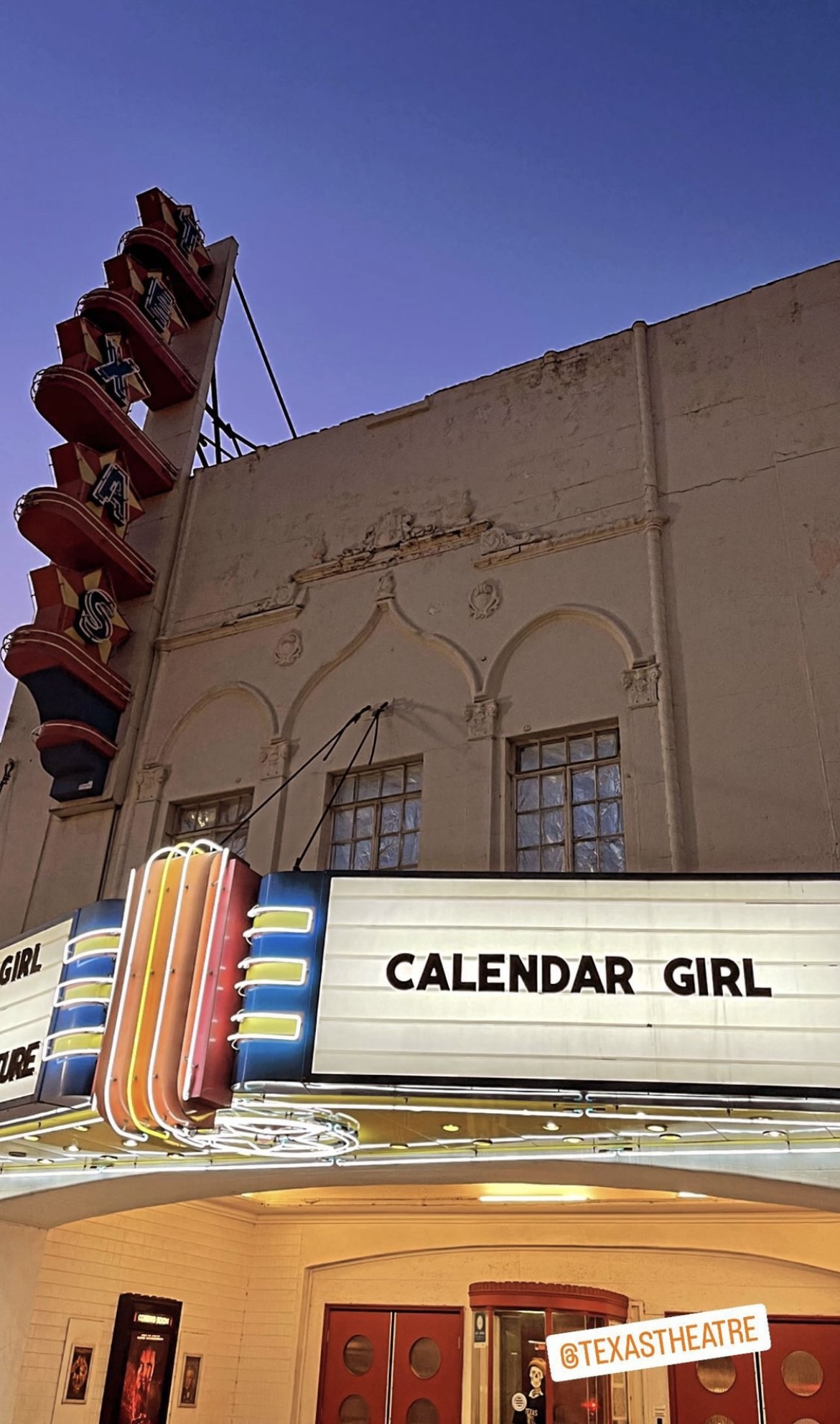 The theater marquee displays 'CALENDAR GIRL' with colorful vertical lights, above a door with red panels, against a beige building.