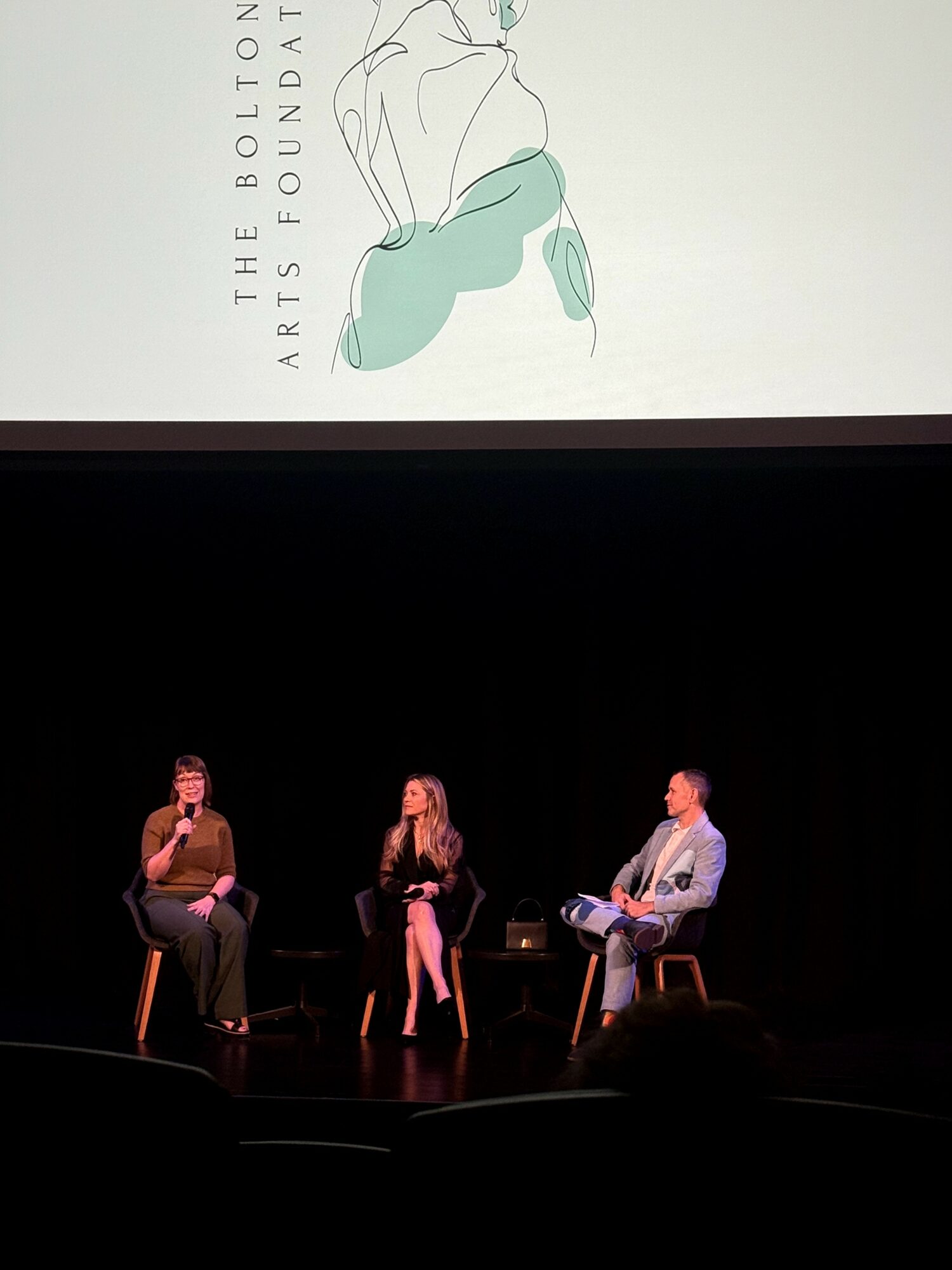 Three people seated on stage in front of a large screen, engaged in a discussion or presentation.