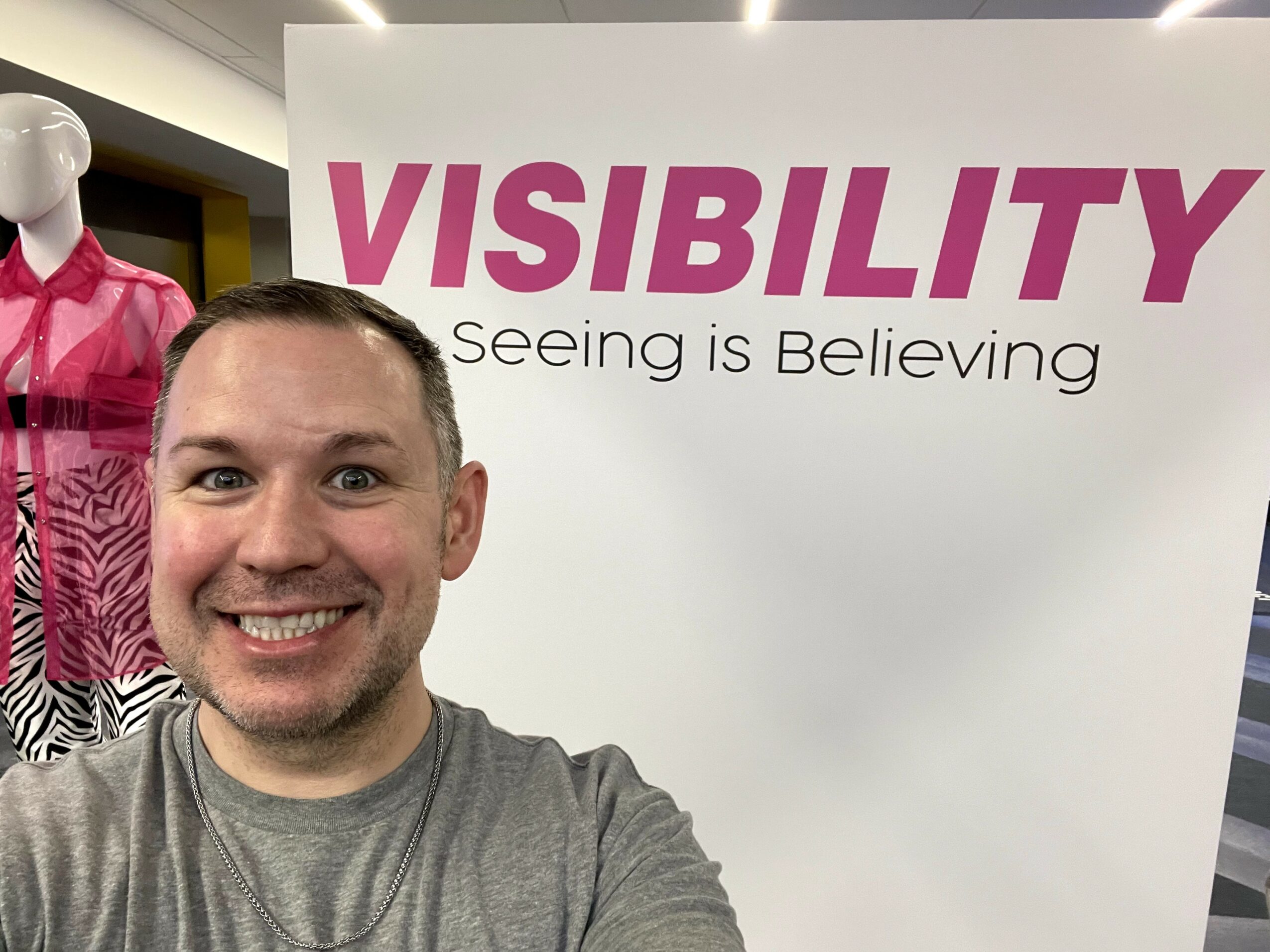 Smiling man in gray shirt standing in front of a sign that says 'VISIBILITY' and 'Seeing is Believing'.