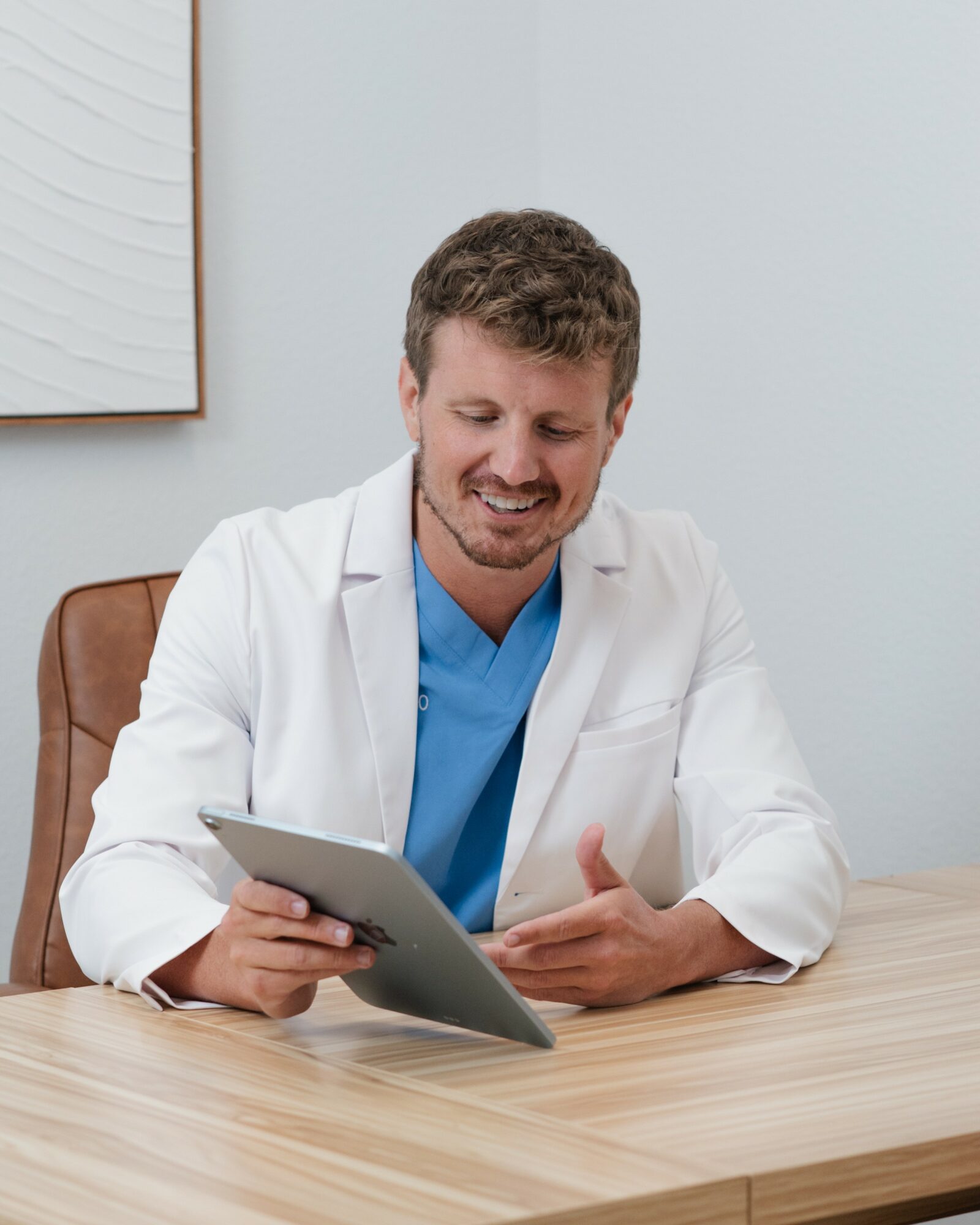 Doctor in white coat and blue scrubs sitting at a desk, smiling while looking at a tablet device.