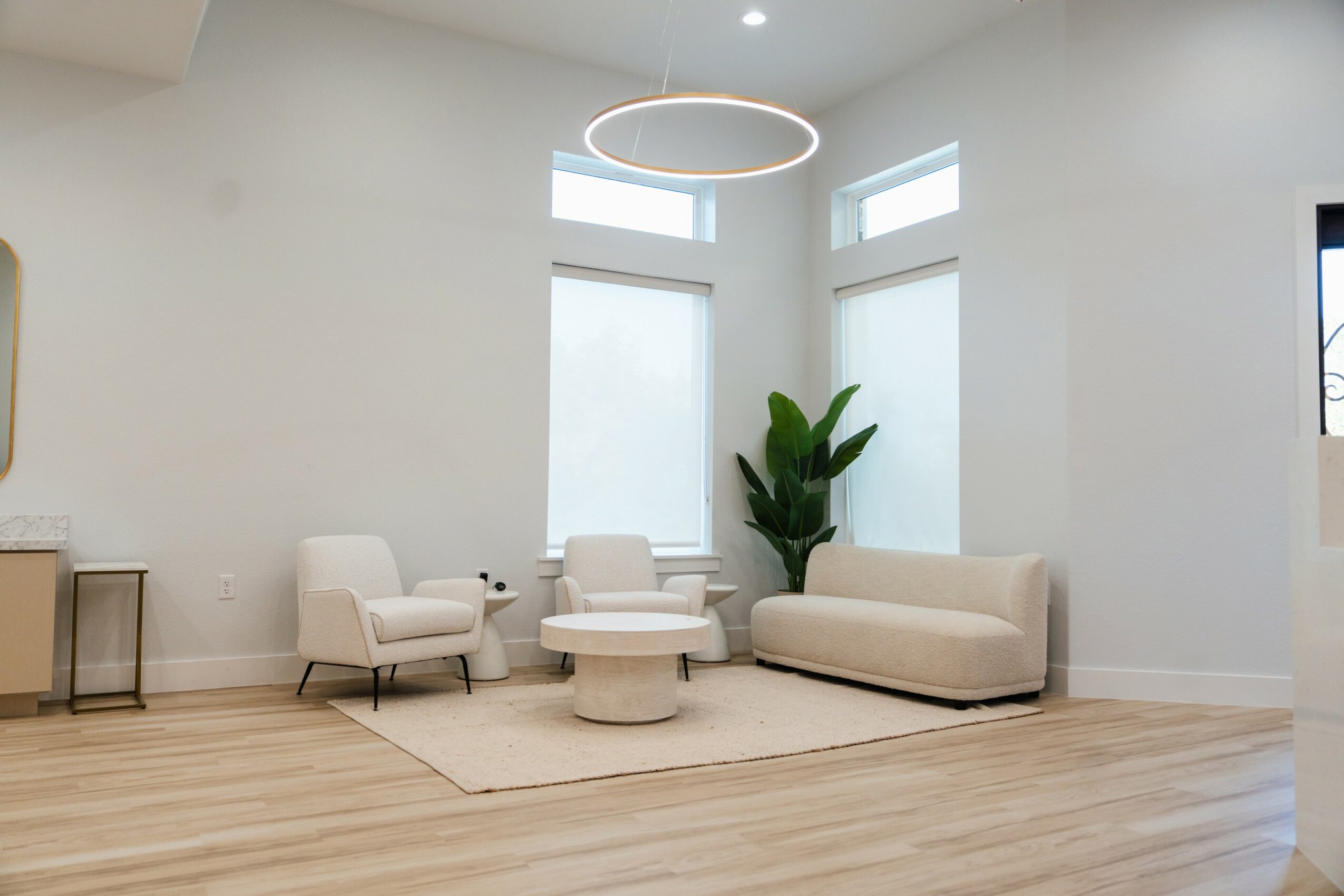 Modern living room with white walls, large windows, a beige sofa, two armchairs, a round coffee table, and a tall plant.