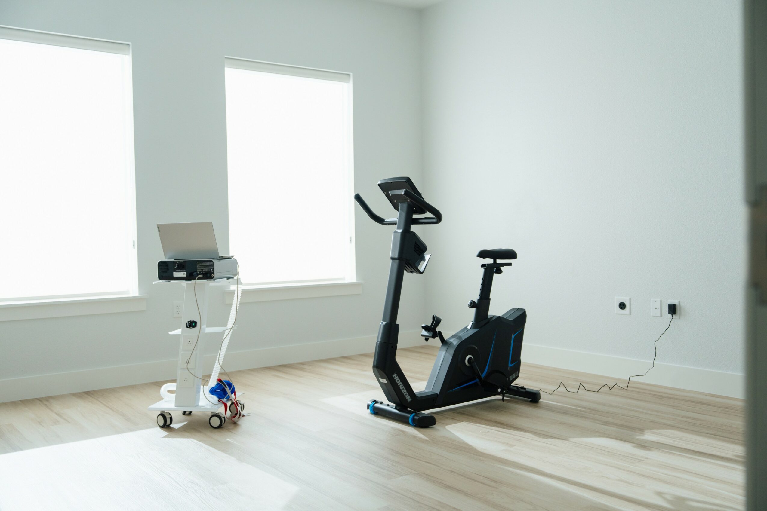 Empty room with two large windows, a stationary exercise bike, and a small white table with a device on top.