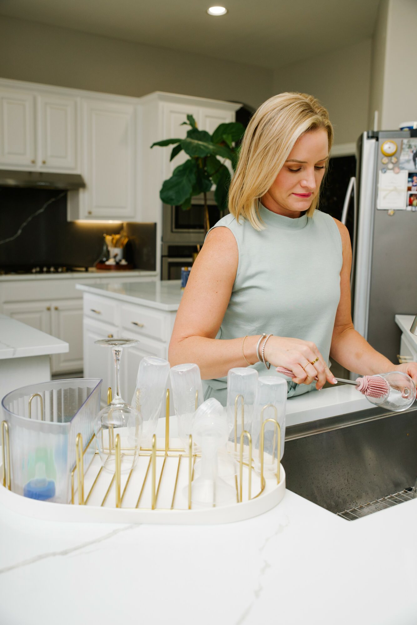 Woman with blonde hair in a sleeveless light blue top preparing food in a modern kitchen, with white cabinets and a large plant in the background.